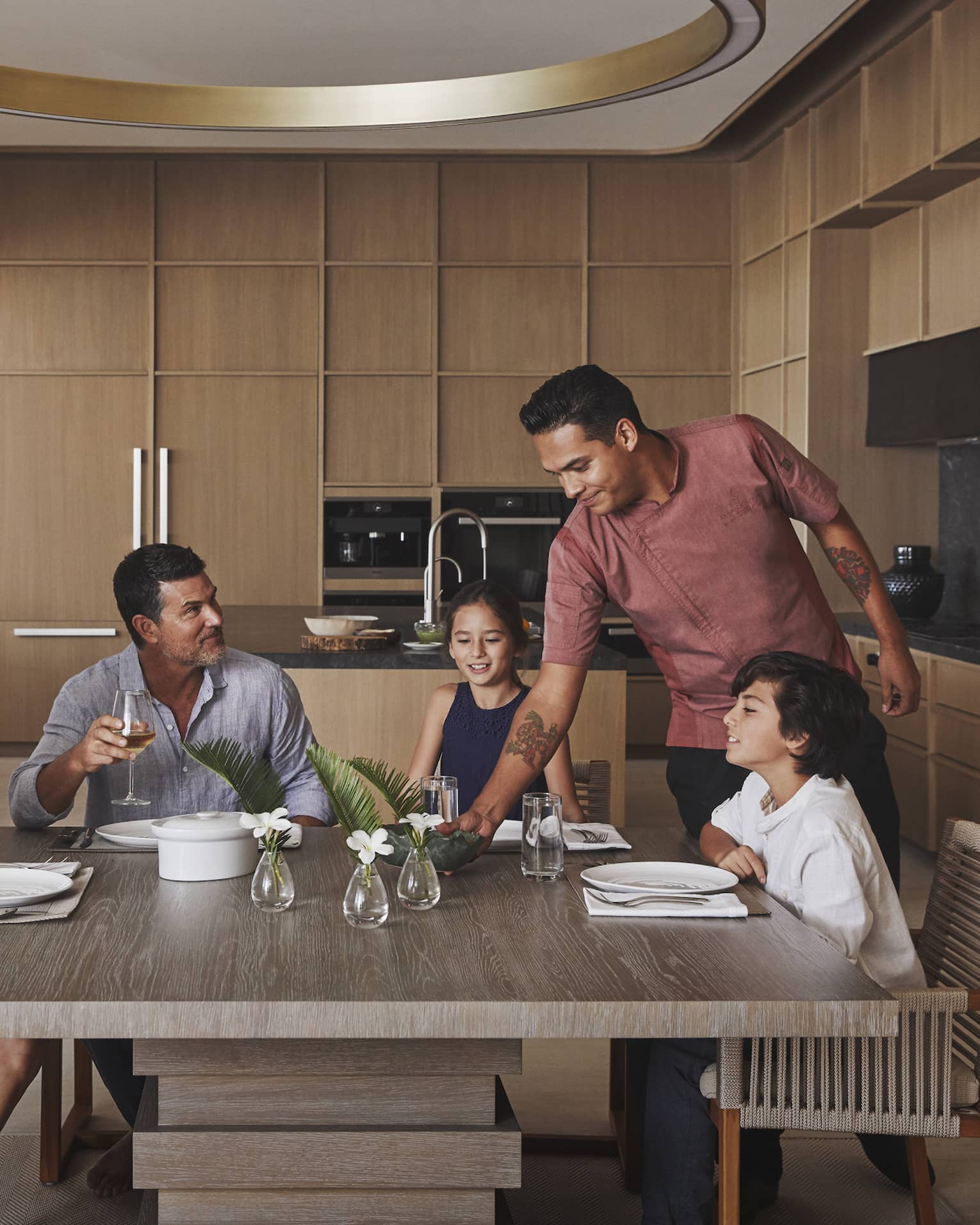 Four guests sitting at the table in a villa while a chef places a dish on the table.