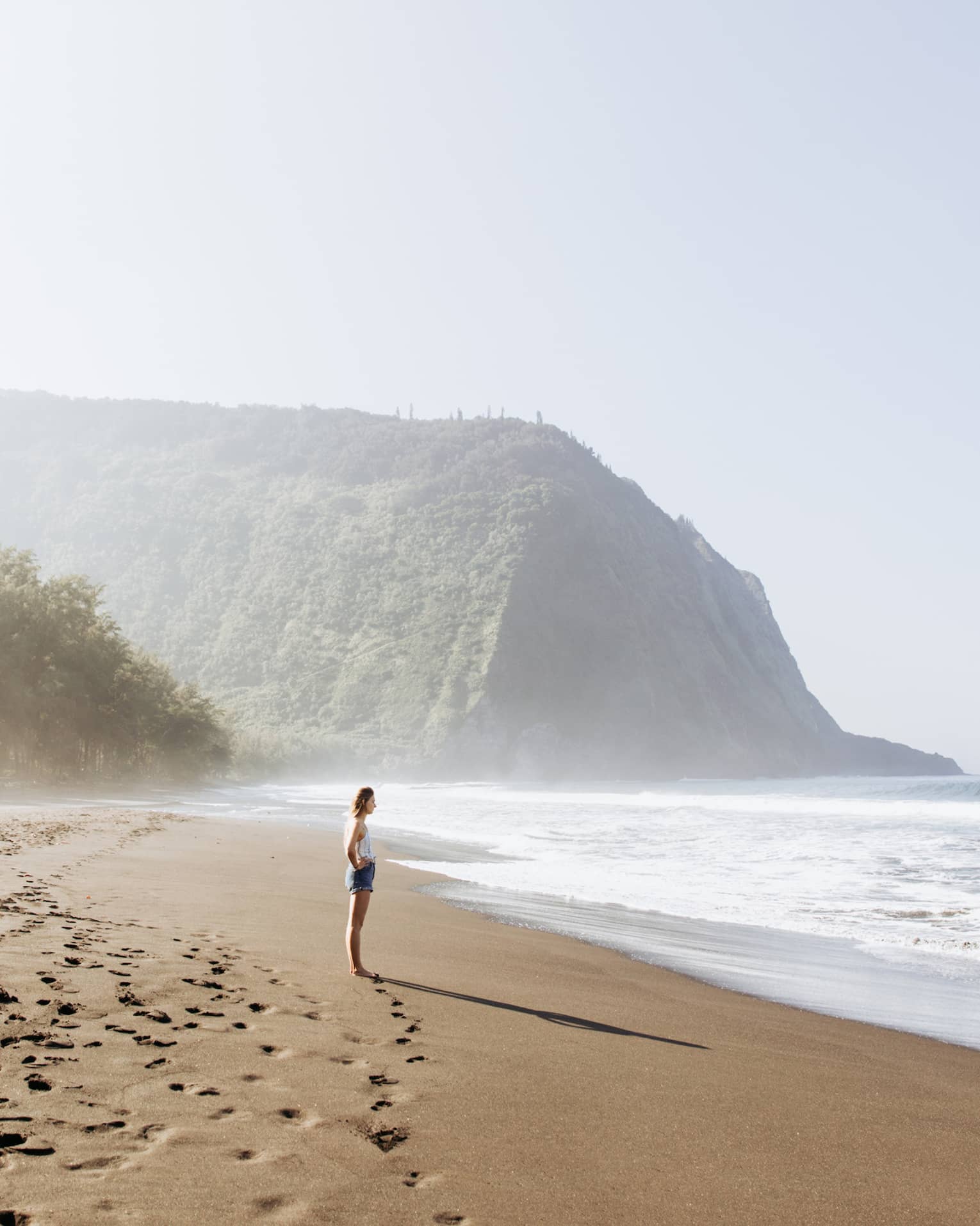 On caramel-coloured sand dotted by footprints, a beachgoer gazes into the ocean; a lush, misty mountain in the background.