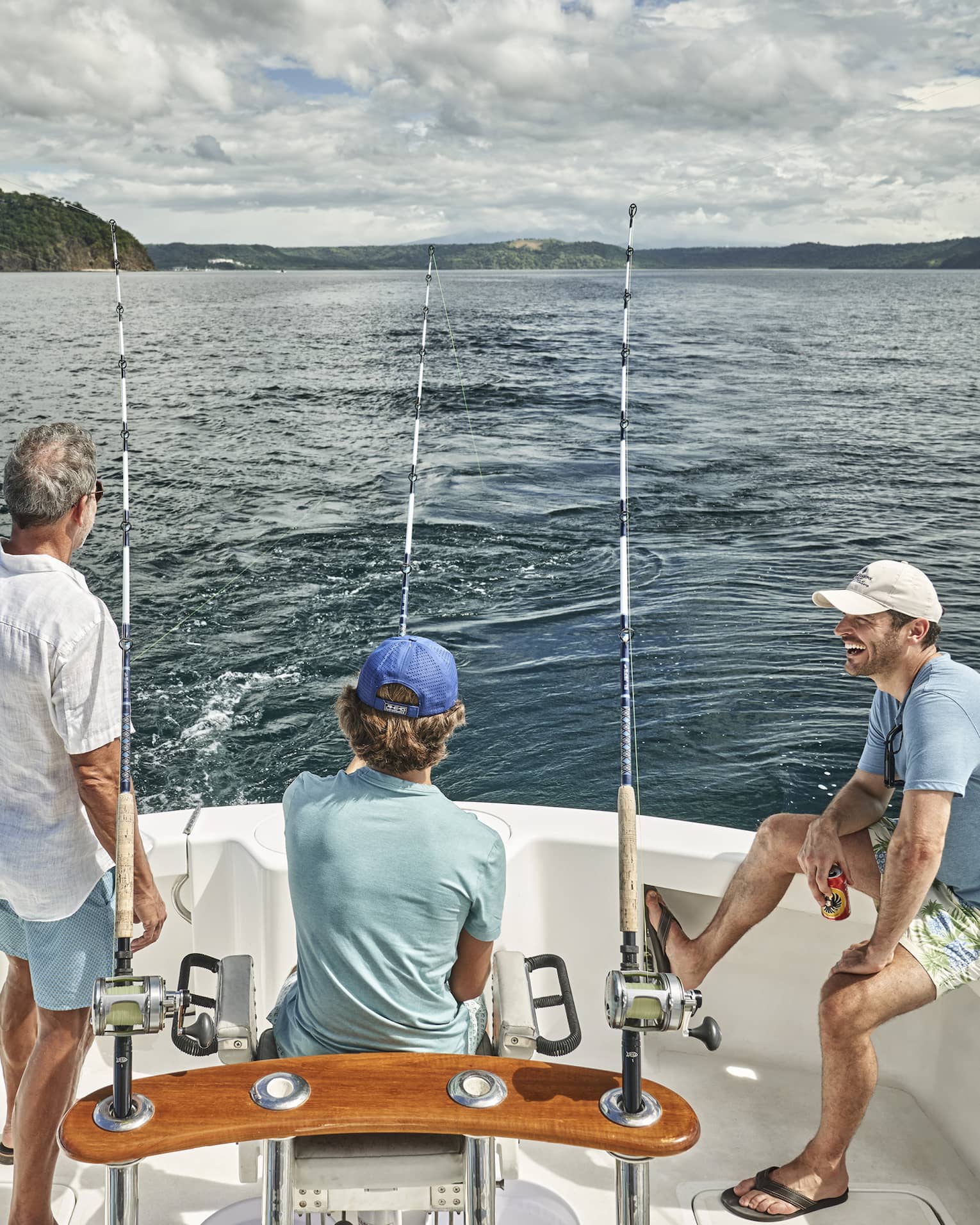 Three men of varying ages sit looking out at the water from the back of a boat outfitted with fishing rods