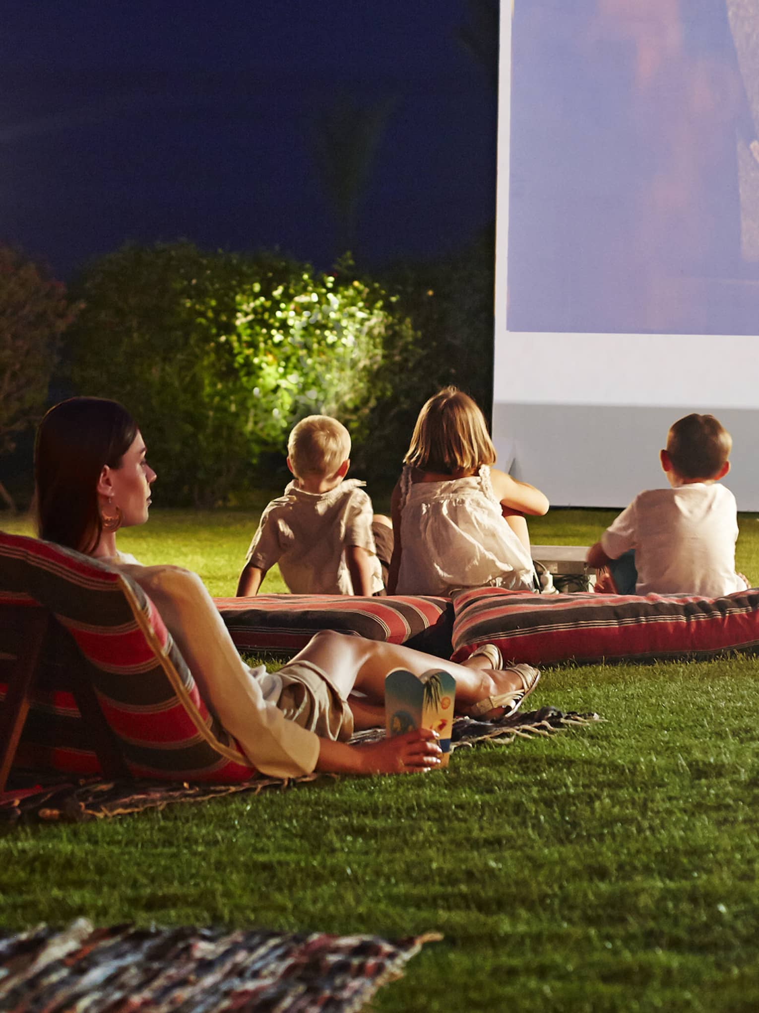 Children, parents lounge on pillows on grass in front of large outdoor movie screen at night