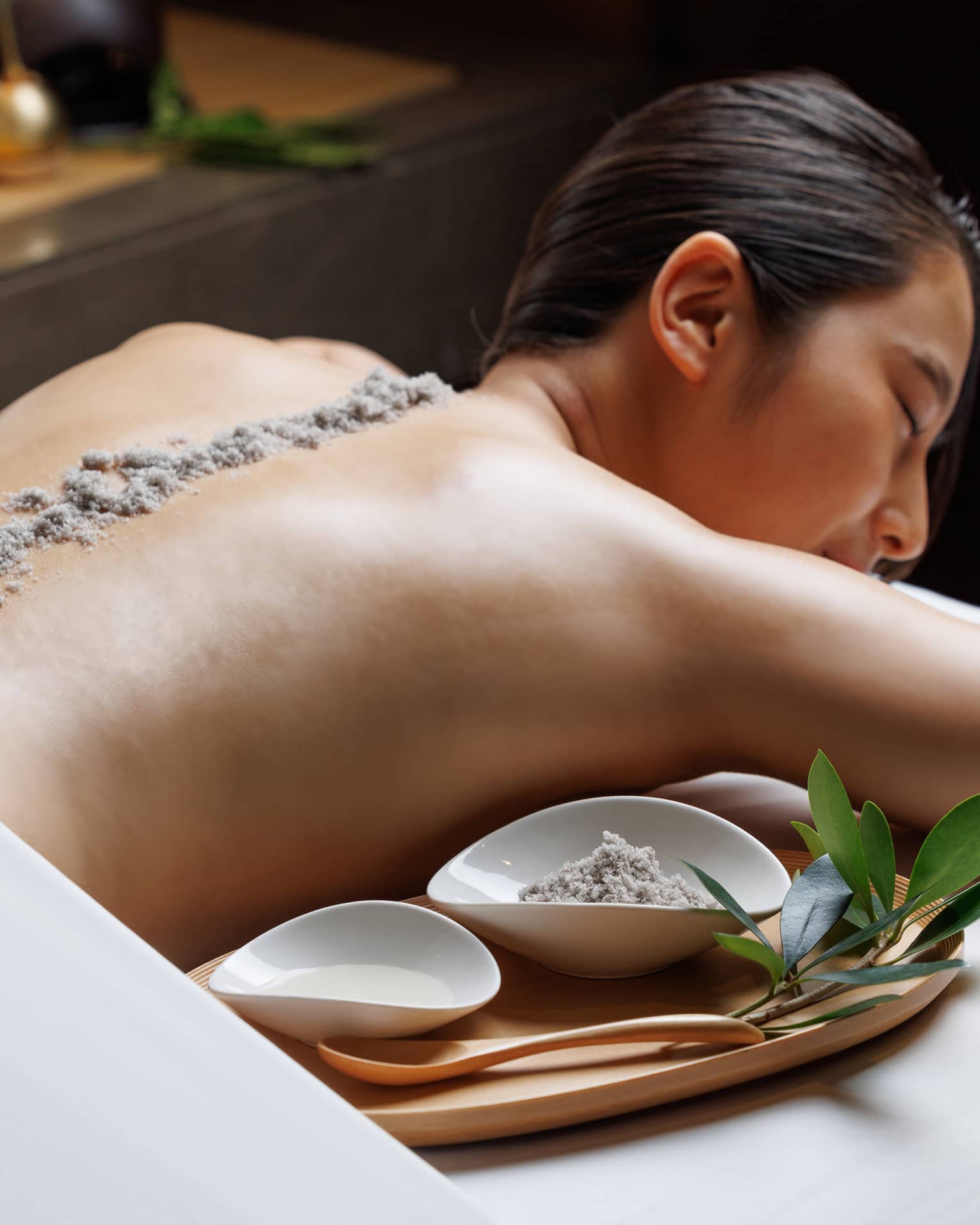 Female guest lies facedown on spa treatment table with line of exfoliating sand running the length of her spine