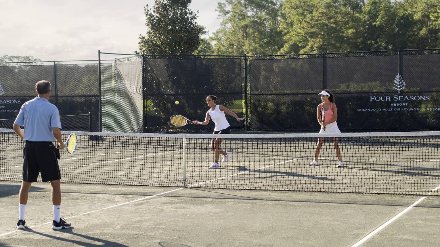 A smiling player prepares to hit the ball while two other players watch from the court while standing in ready position.