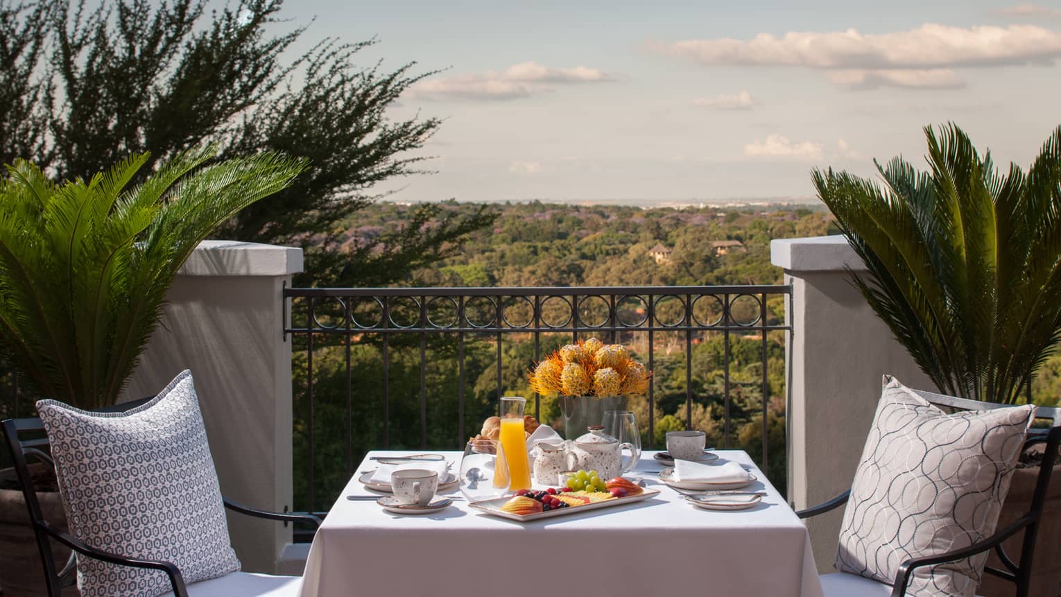 Dining table with breakfast dishes, juice, fresh fruit against iron balcony overlooking canopy of trees