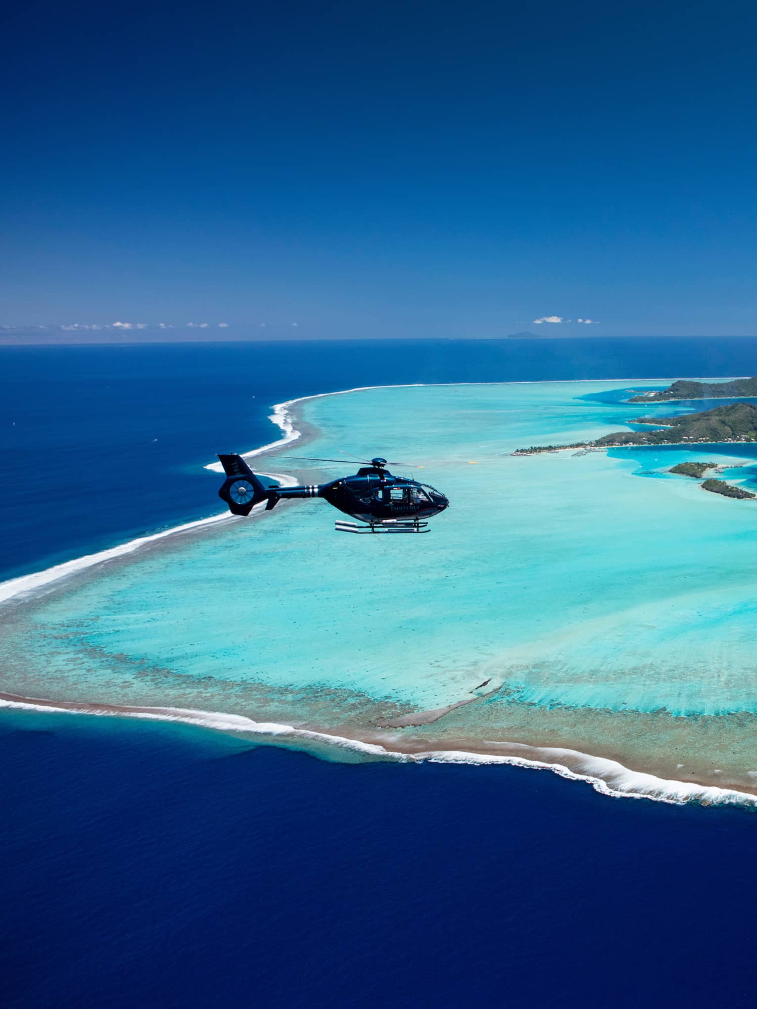 Aerial view of a helicopter flying over Bora Bora and the distinctive pastel blue lagoon that surrounds the islands.
