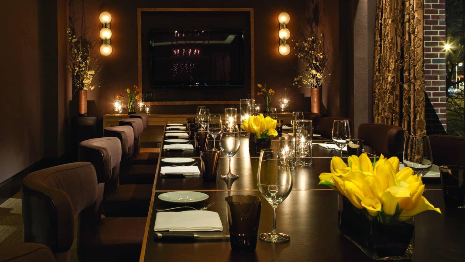 Long private dining table with plush chairs set with wine glasses, yellow flowers, candles, low lights on walls