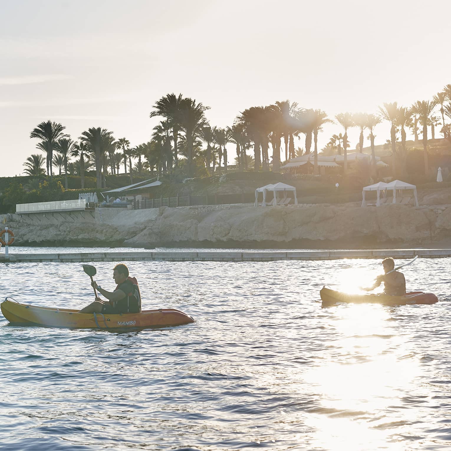 Two kayakers in a blaze of sun paddle near a resort dock and rock-faced shore lined with white beach cabanas and palm trees.