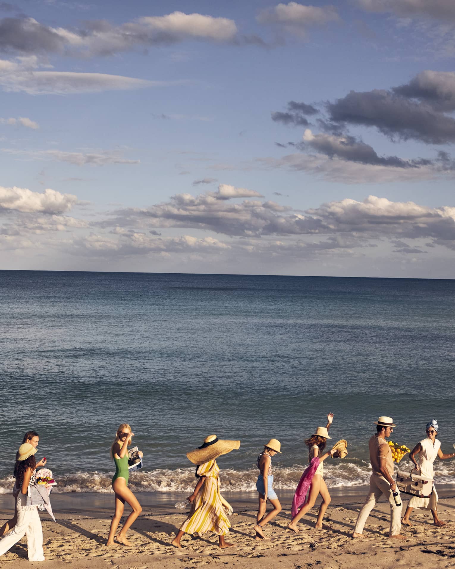 A group of people walking along a sandy beach by the ocean, wearing summer clothing and hats, with a backdrop of a calm sea and partly cloudy sky.