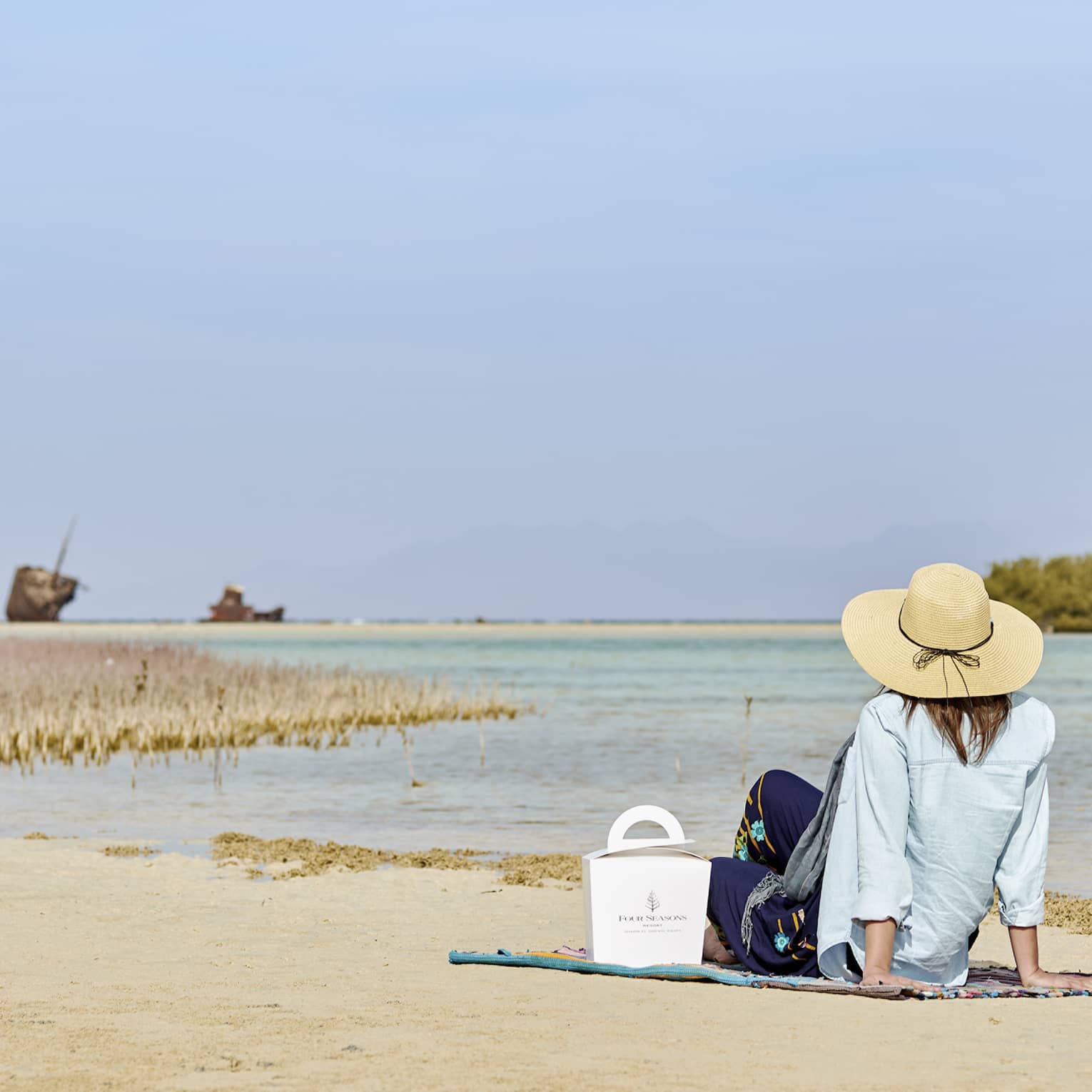 A guest in a sunhat, a Four Seasons food container beside them, sits on a beach by the water's edge, gazing at the horizon.