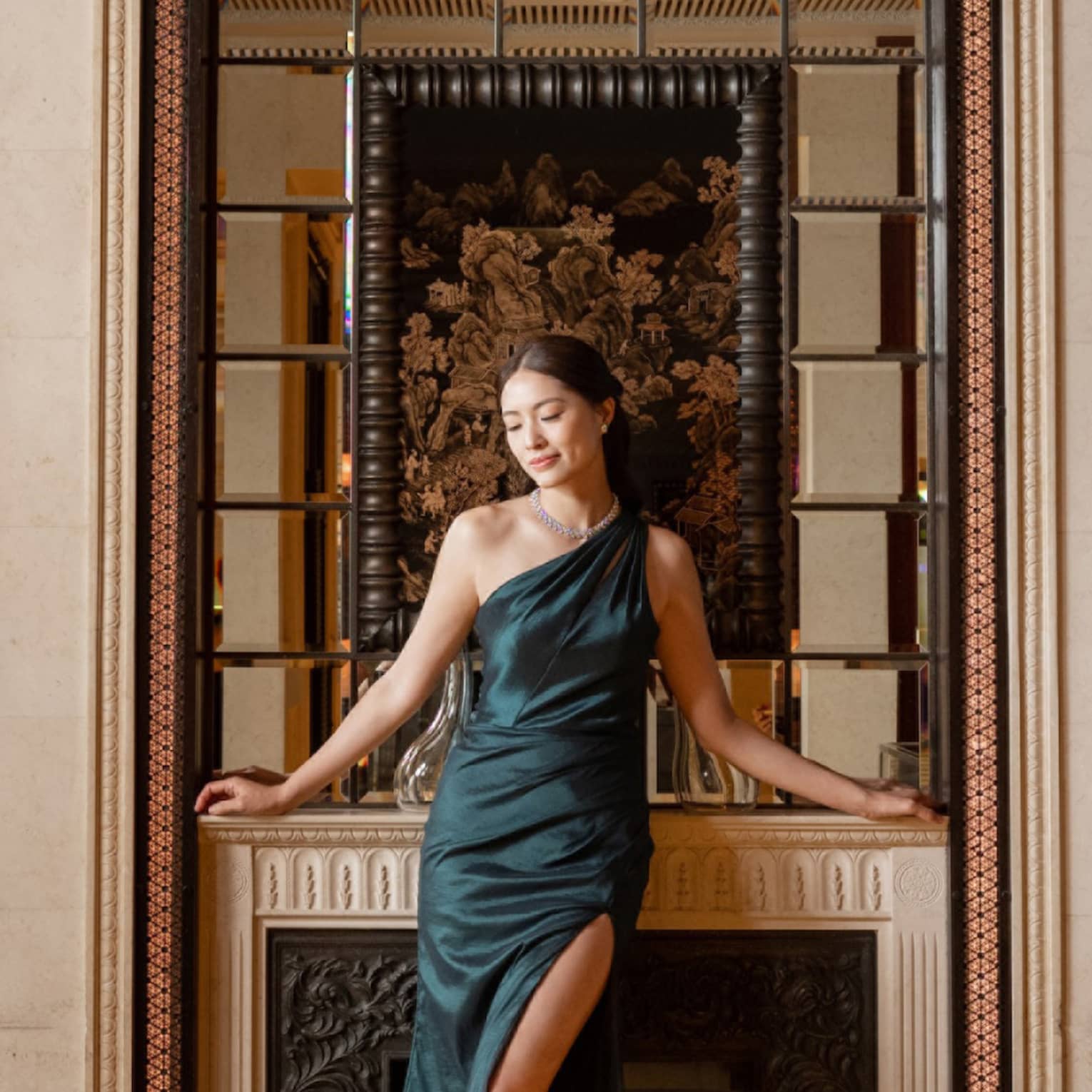 A formally dressed guest posing in front of an elegant fireplace with bottles of wine in tabletop glass cases on either side.