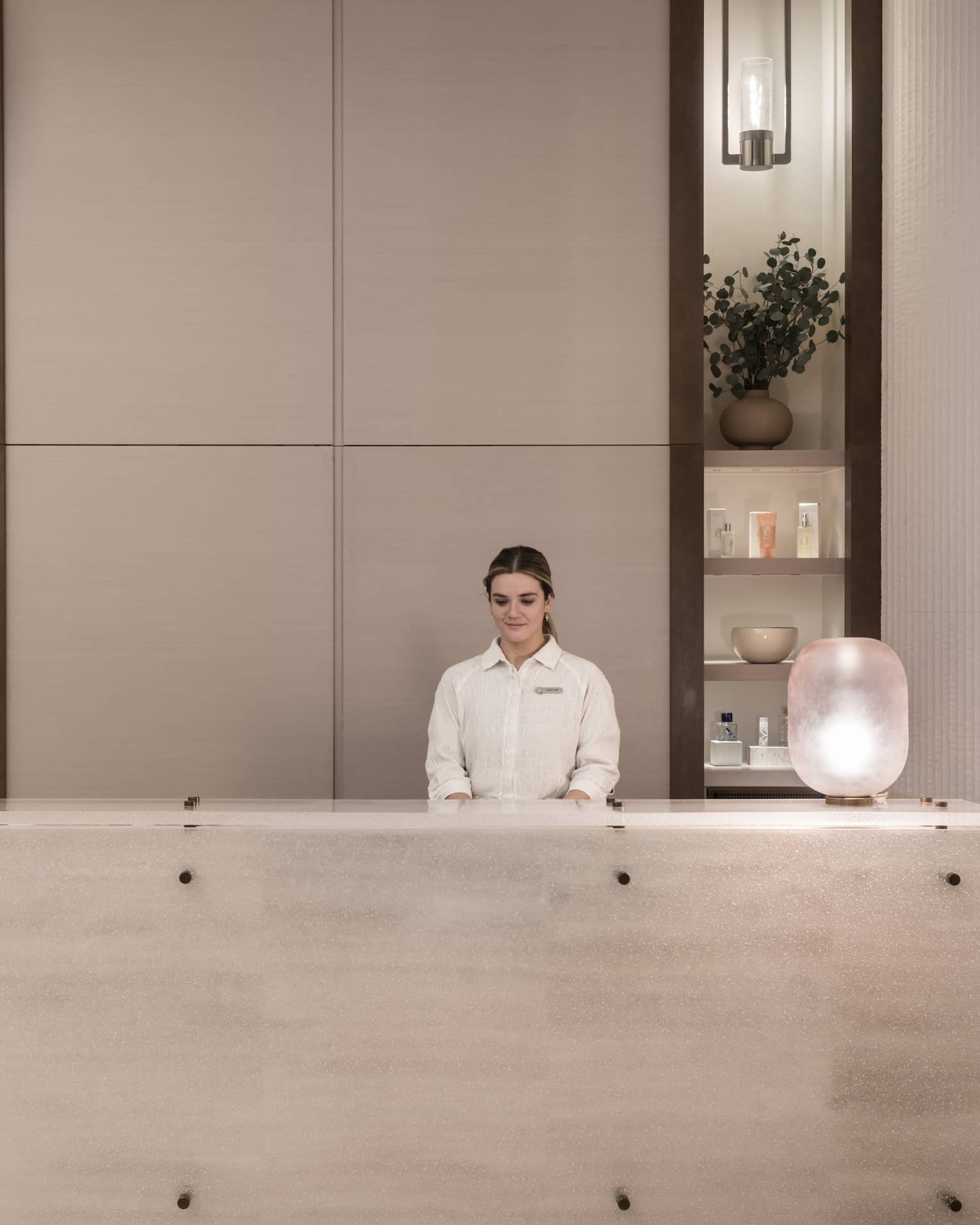 Receptionist in white shirt stands behind white marble spa reception desk with shelving display in backdrop