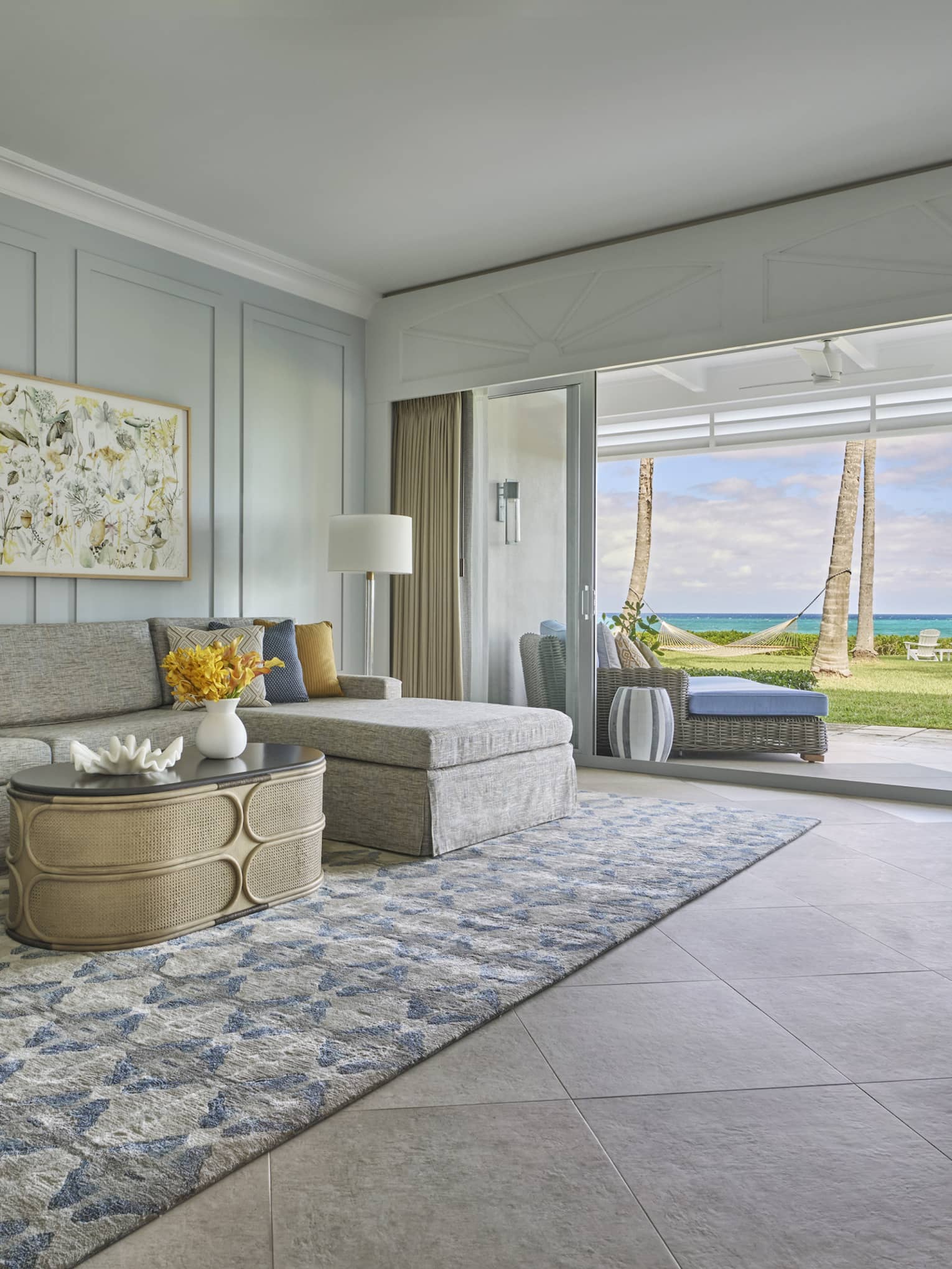 Living room with a gray sectional sofa, blue armchair, patterned rug and glass doors opening to an ocean view with palm trees