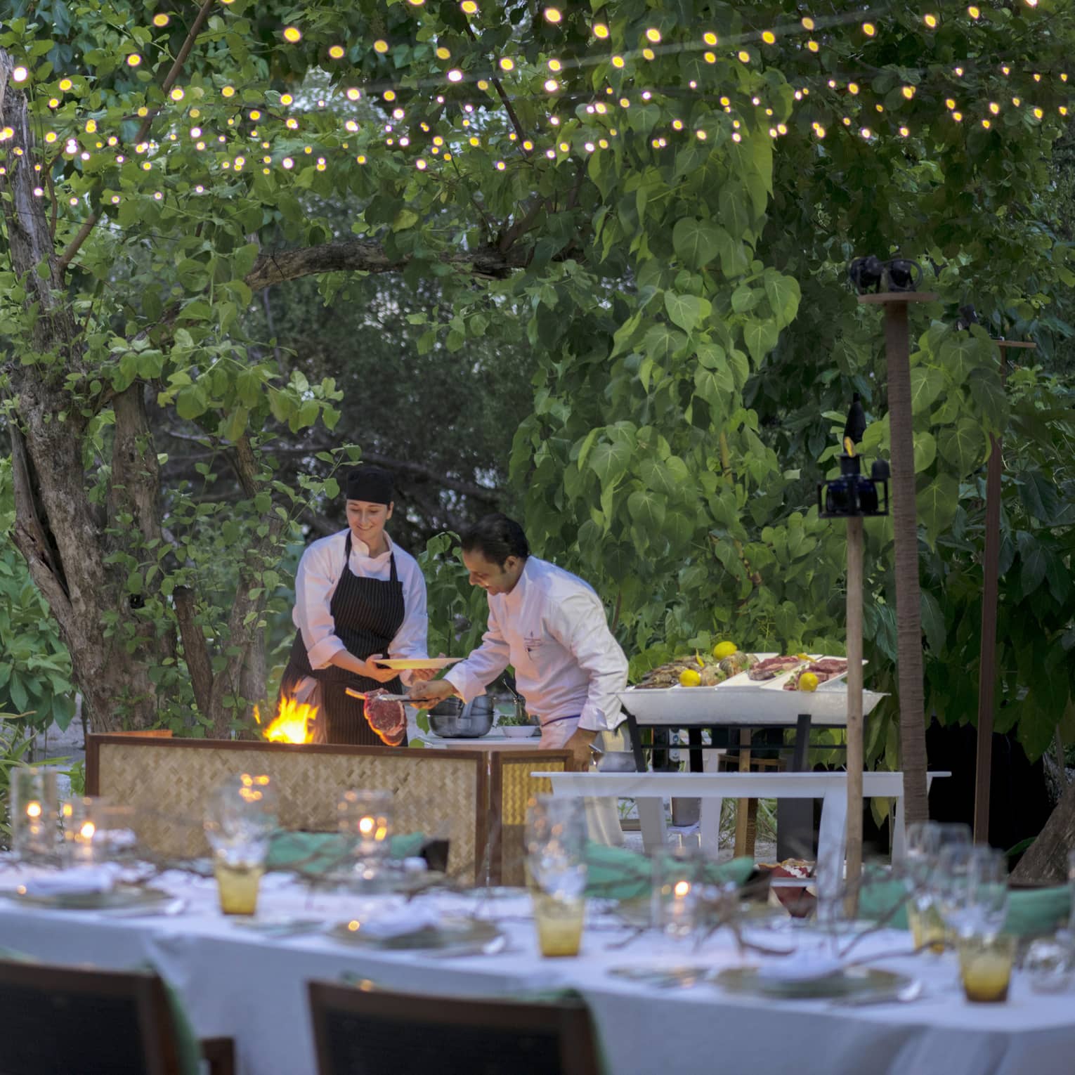 Chefs put large steak on flaming grill by long outdoor dining table, patio lights at night