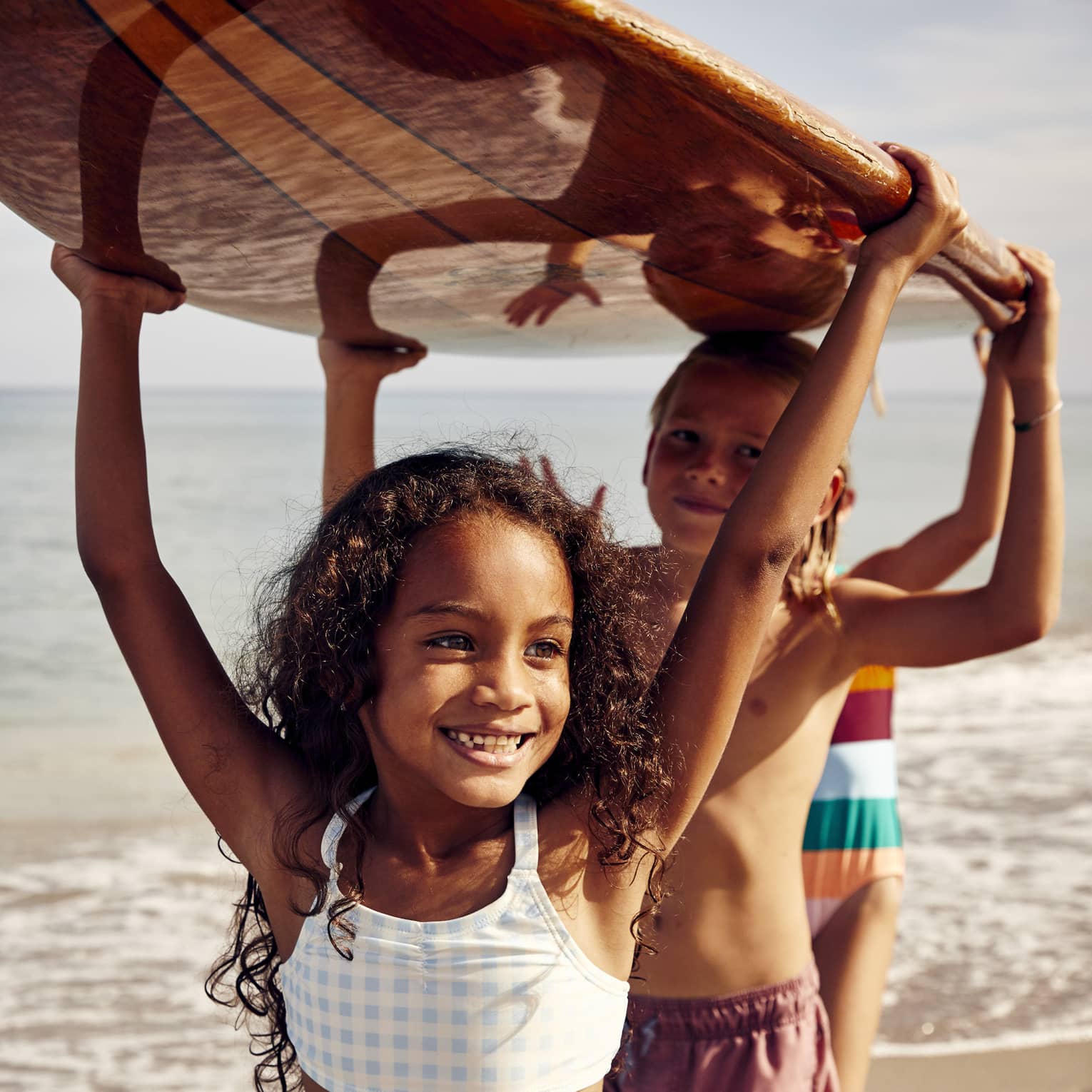 Three young children carry a surfboard on the beach, smiling and enjoying the sunny day. The ocean is in the background.