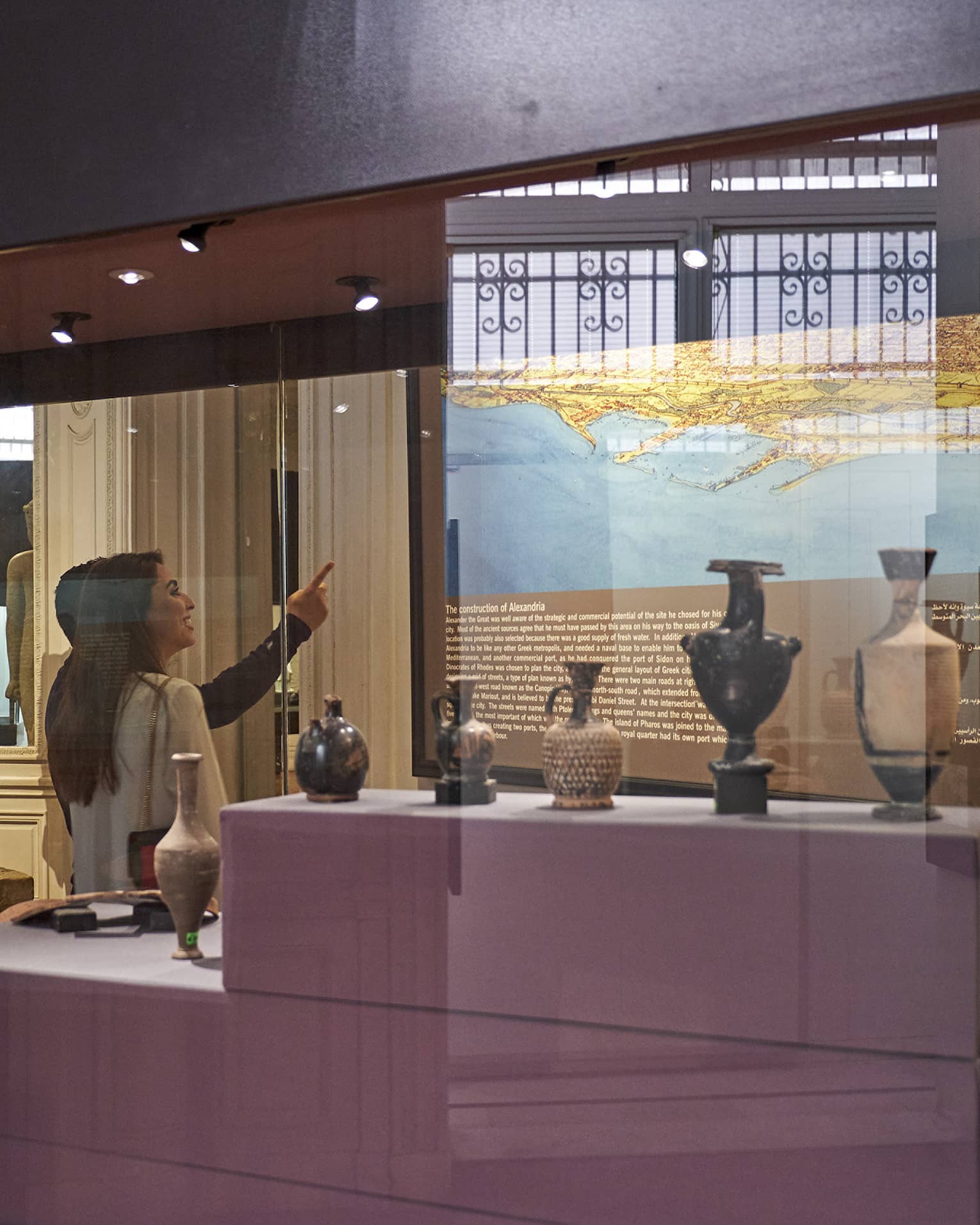 A visitor in a museum points up at a historical map lit up on a wall, behind them a display case with various vases and jugs.