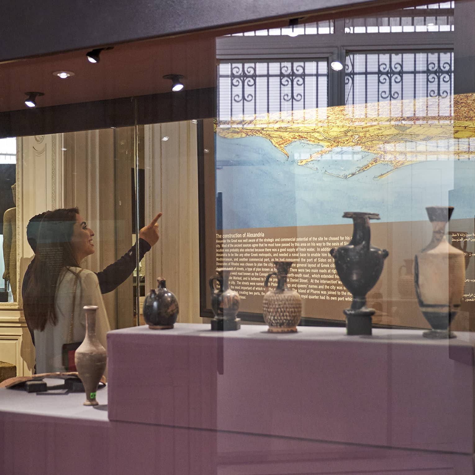 A visitor in a museum points up at a historical map lit up on a wall, behind them a display case with various vases and jugs.