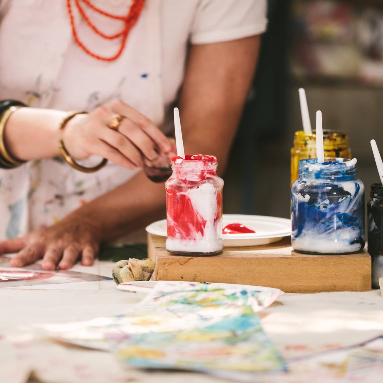 An artist using mason jars filled with red, blue, yellow and black paint to paint on a table covered in paper paintings.