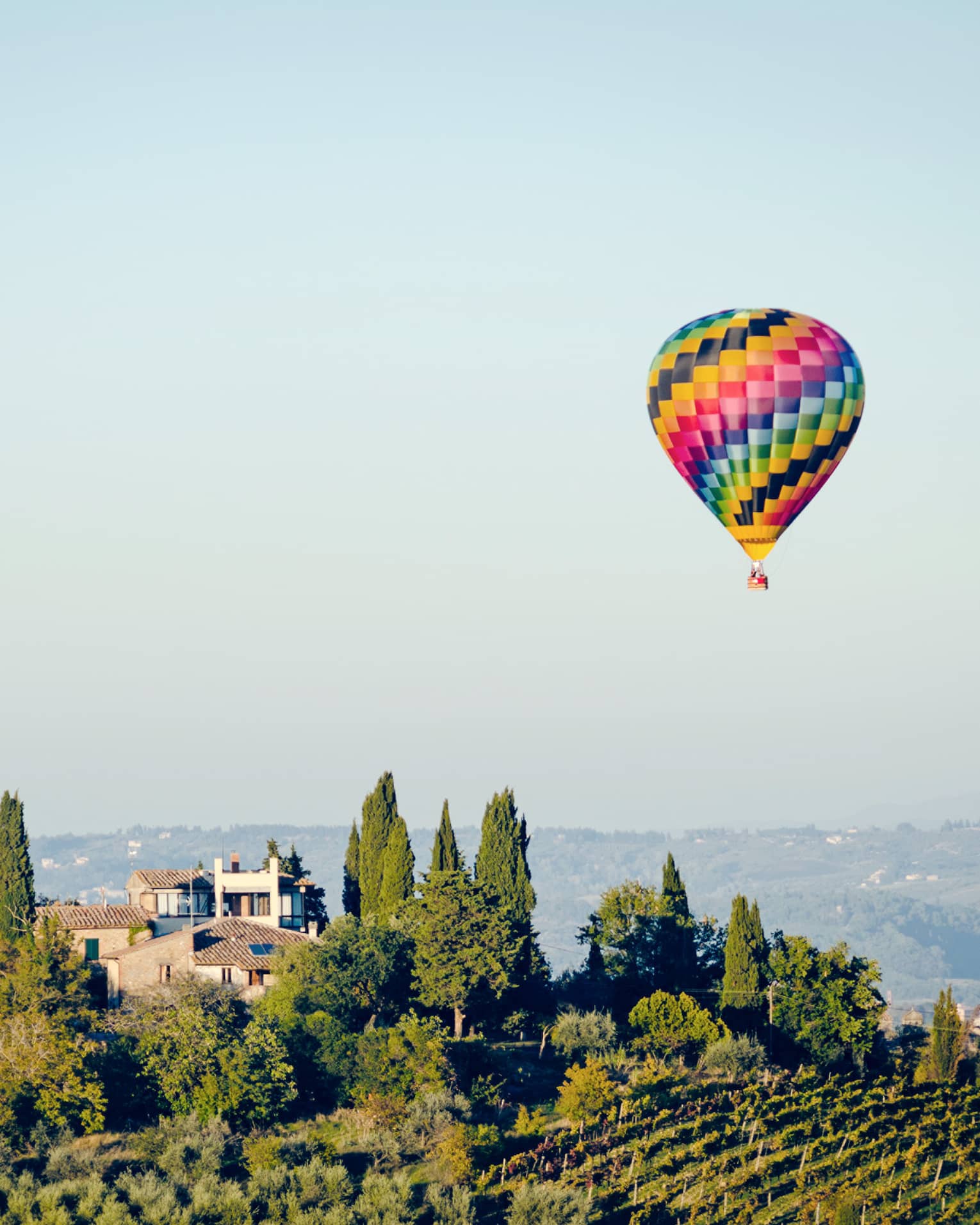 Colourful hot air balloon floating above a landscape with trees and houses, set against a clear blue sky.		