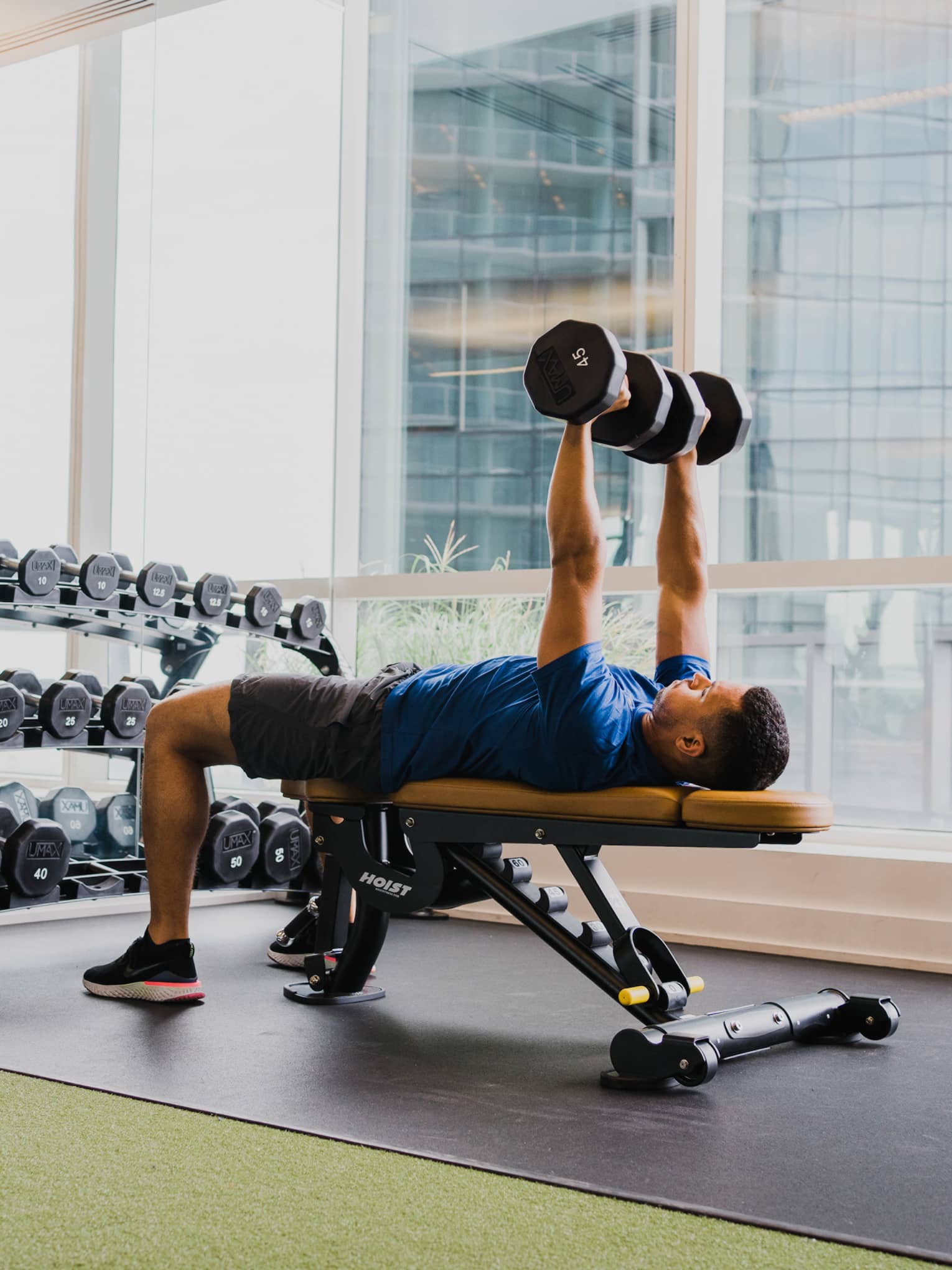 A man working out with free weights in a clean fitness room with large windows.