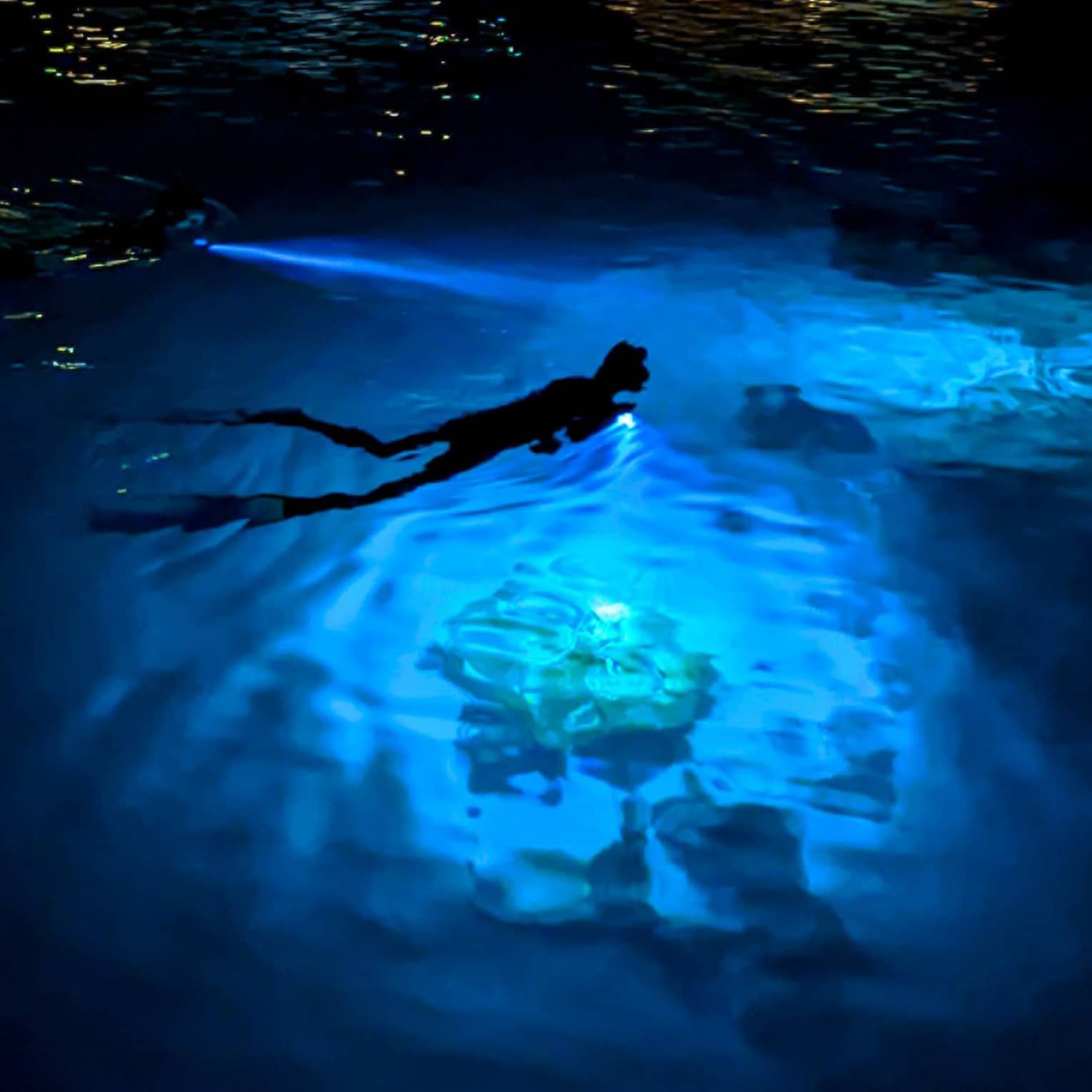 Aerial view of a night-time snorkeller with an underwater flashlight gliding through a bright spot in the dark ocean water.