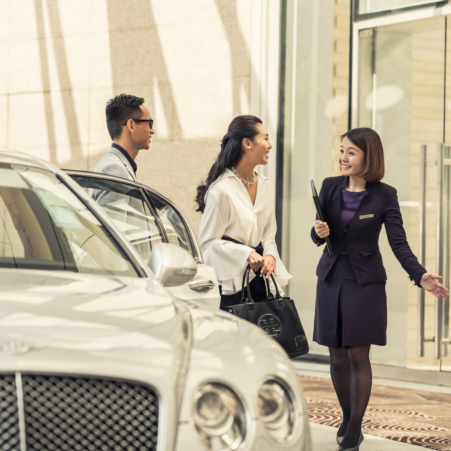 A person in a dark suit greets a couple near a luxury white car at the hotel entrance. The couple appears enthusiastic and engaged in conversation.