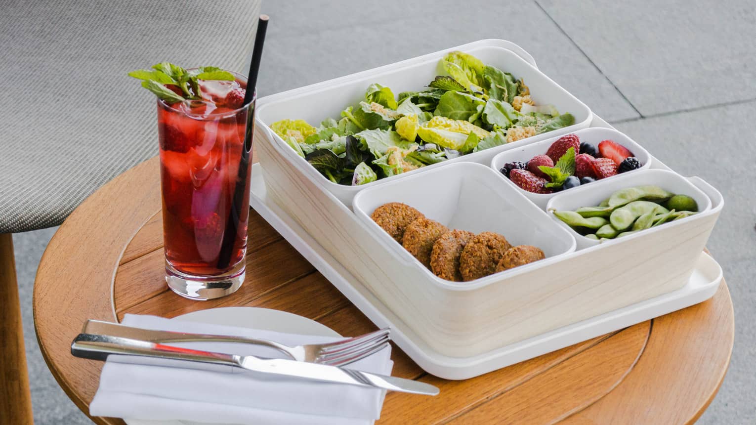 A white bento box filled with an assortment of colorful foods sits next to a red-colored beverage in a tall glass, both on a small round wooden table