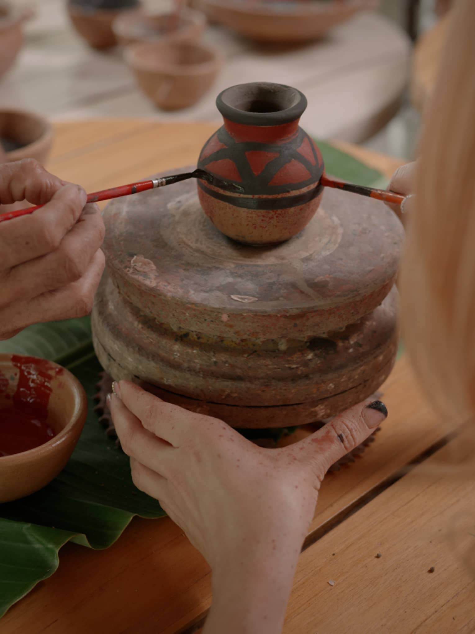 Two sets of hands using thin paintbrushes to decorate a small ceramic vase on a pottery wheel amid pots of paint and pigment.