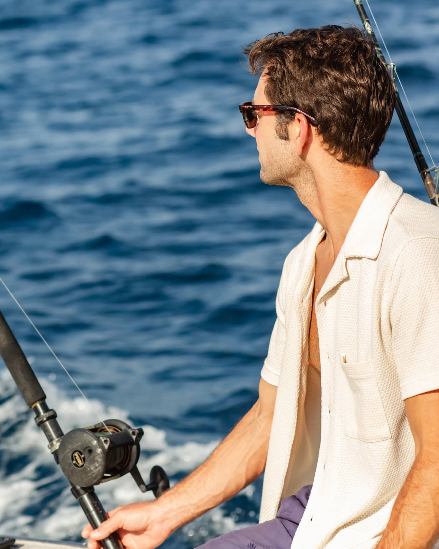Close-up of a guest wearing sunglasses, sitting beside two offshore fishing rods and looking out towards dark blue waves.