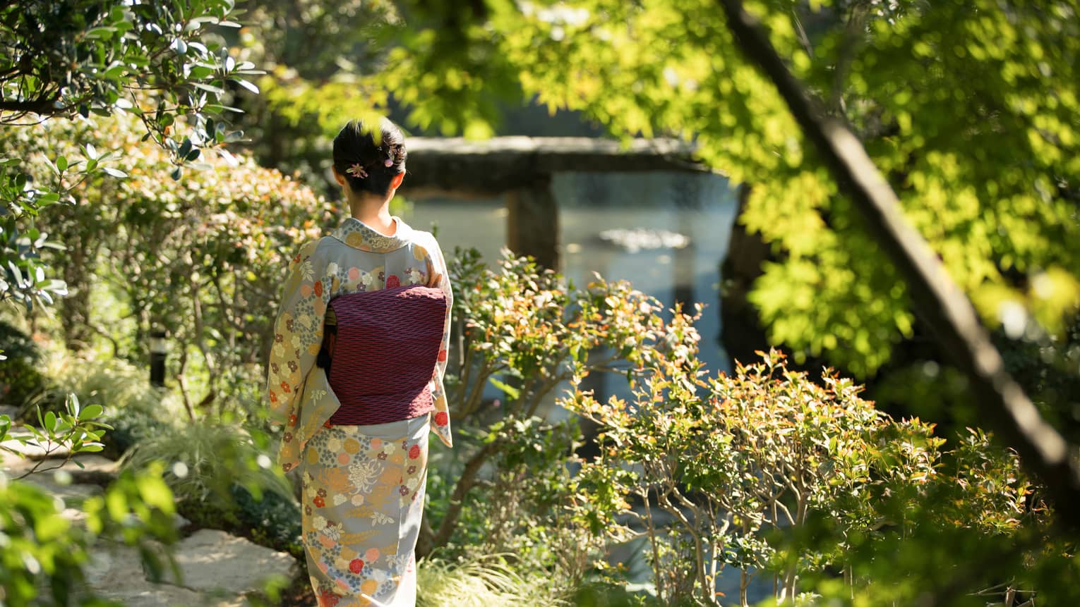 Amid bushes and trees, rear view of a person in a grey kimono with beige and orange flowers and a black and pink obi cushion.