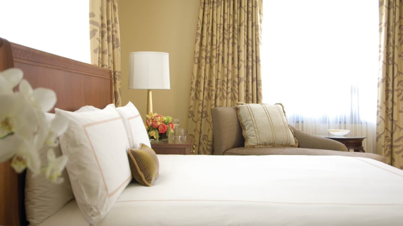 Deluxe City-View Room white orchids, side of bed, pillows against headboard, armchair, window in background