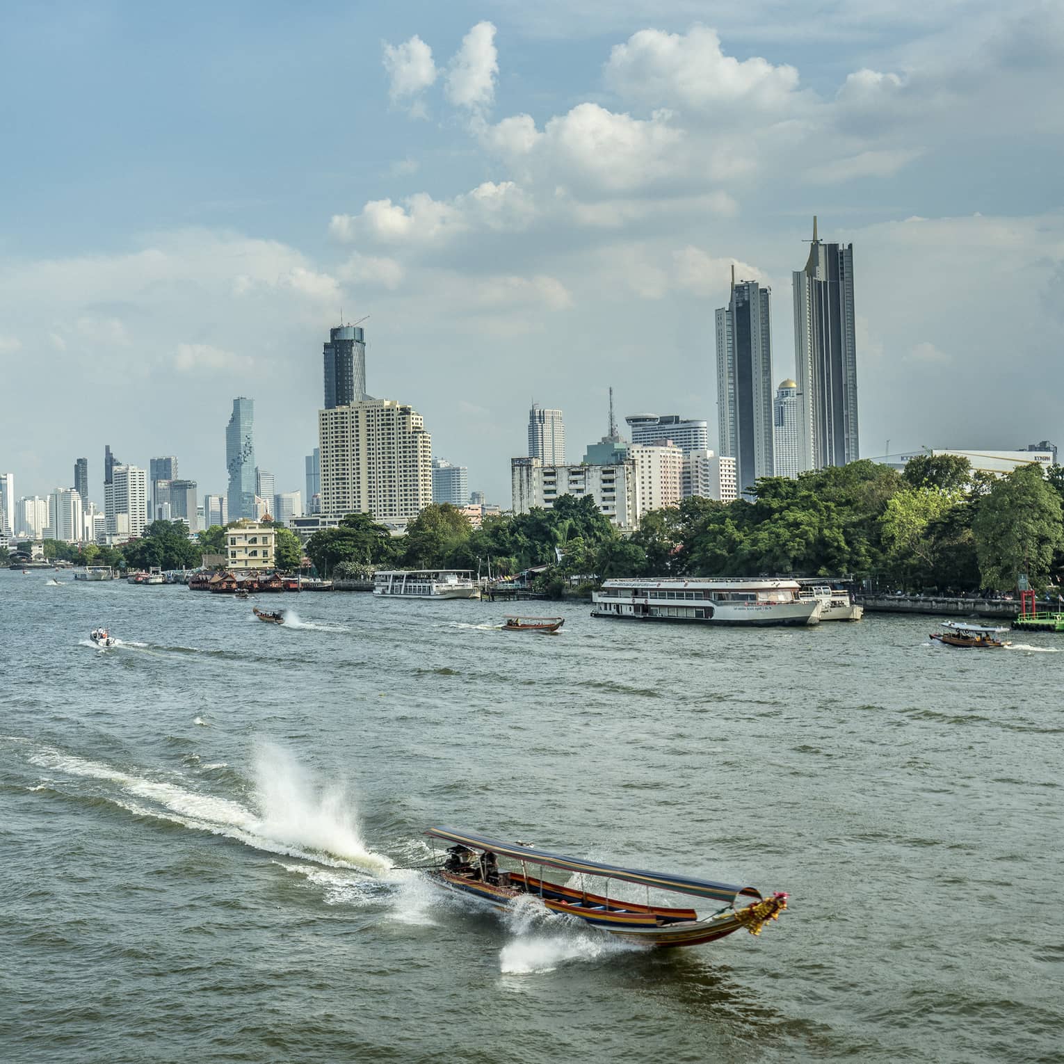 Longtail boat heading into foreground away from Bangkok skyline, leaving trail behind and surrounded by tall, splashing wake.