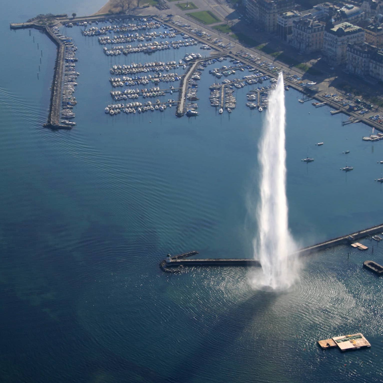 Overhead view of dozens of boats docked at a large marina featuring a tall geiser shooting water high into the air. 