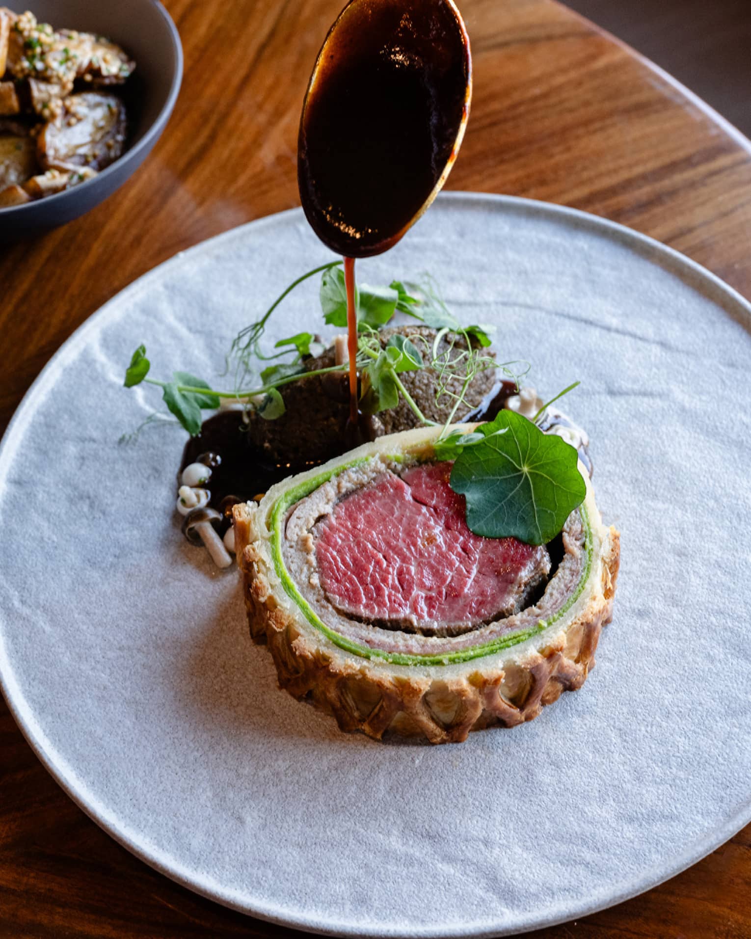 Slice of beef Wellington plated on a white dish with greens on the side plus a bowl of sliced vegetables and a bowl of mushrooms