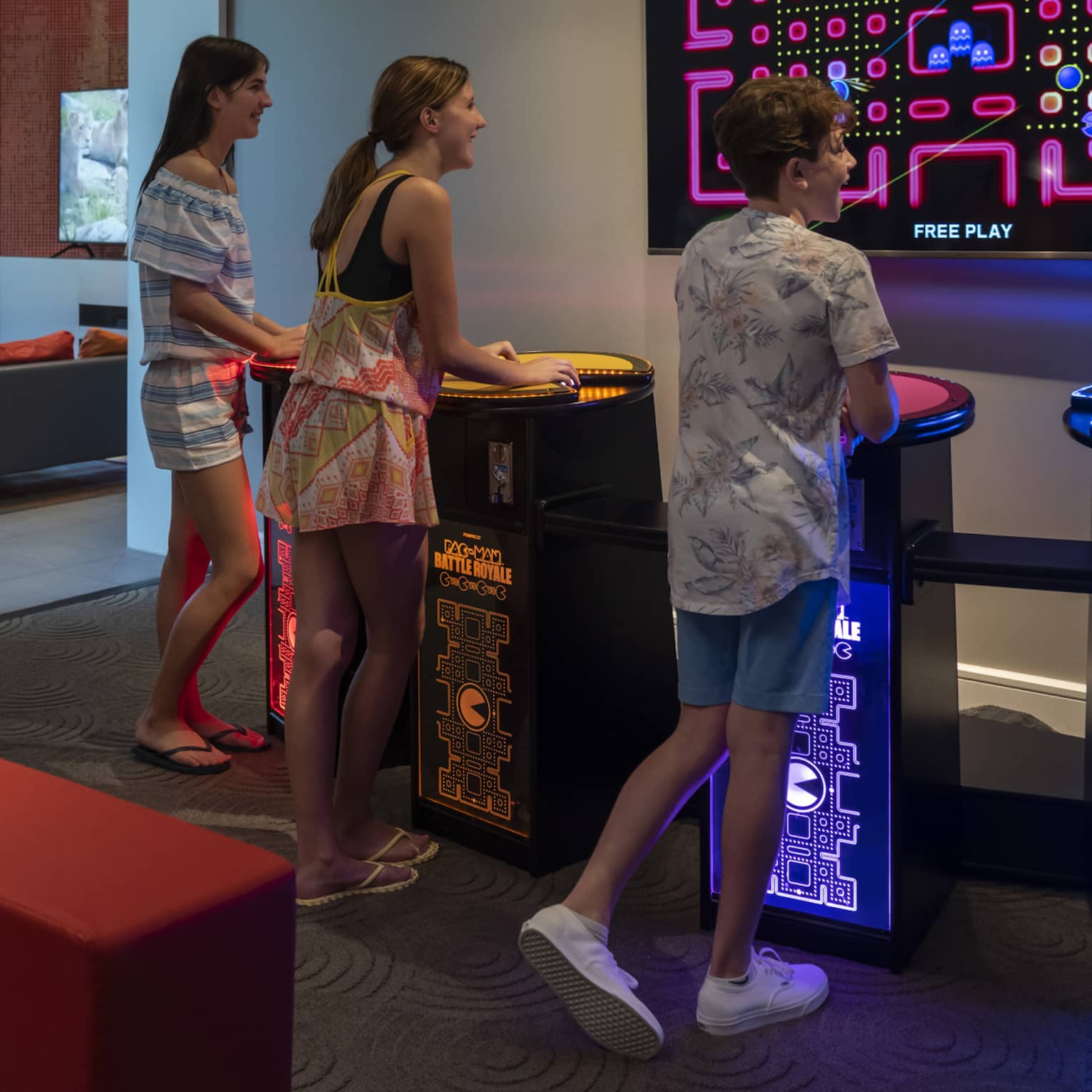 Four smiling teenagers stand at multi-coloured video-game pedestals facing a large TV screen in a dimly-lit games room.