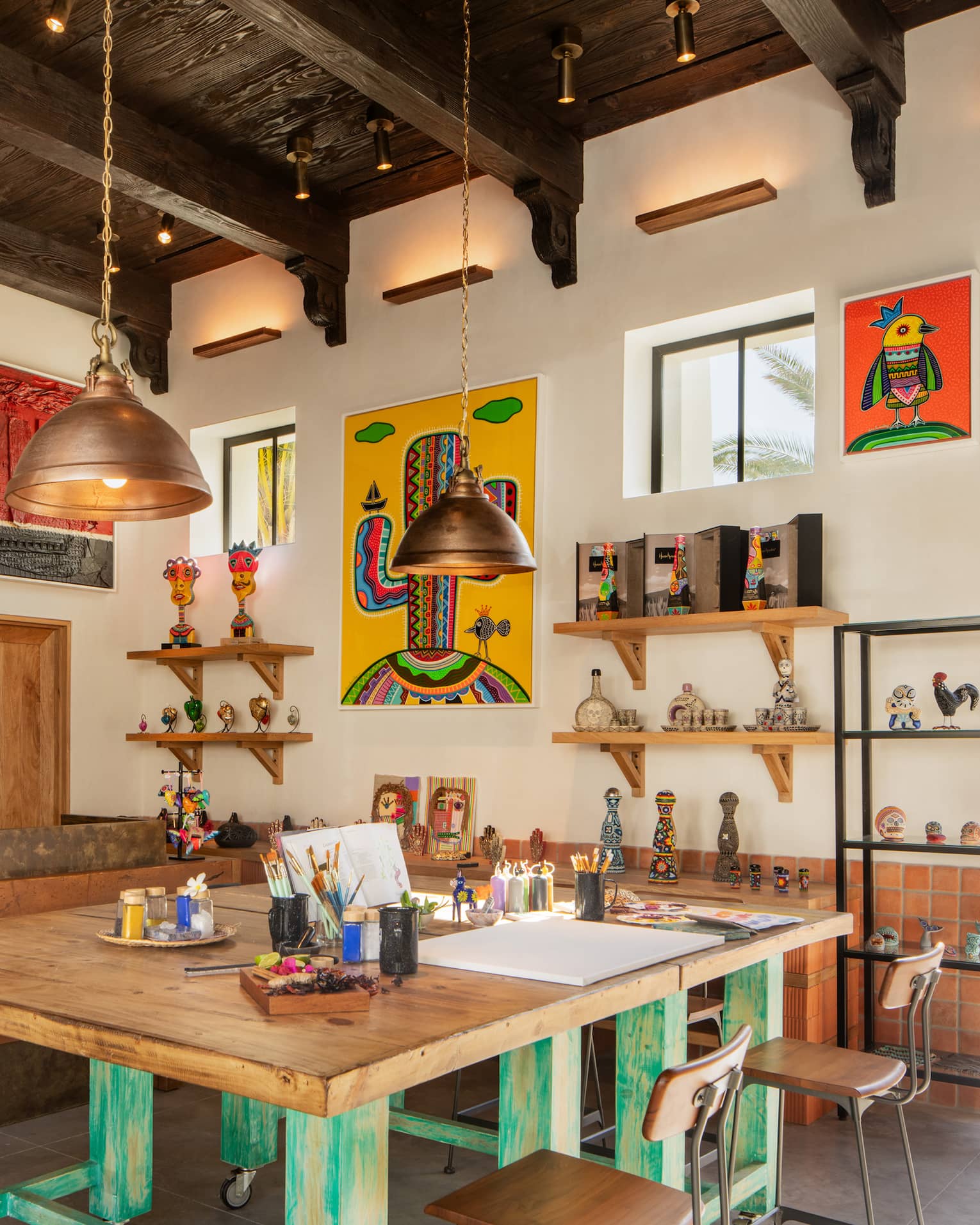 A bright, airy art studio with a large square wooden worktable, shelves of knick-knacks and colourful paintings on the walls.