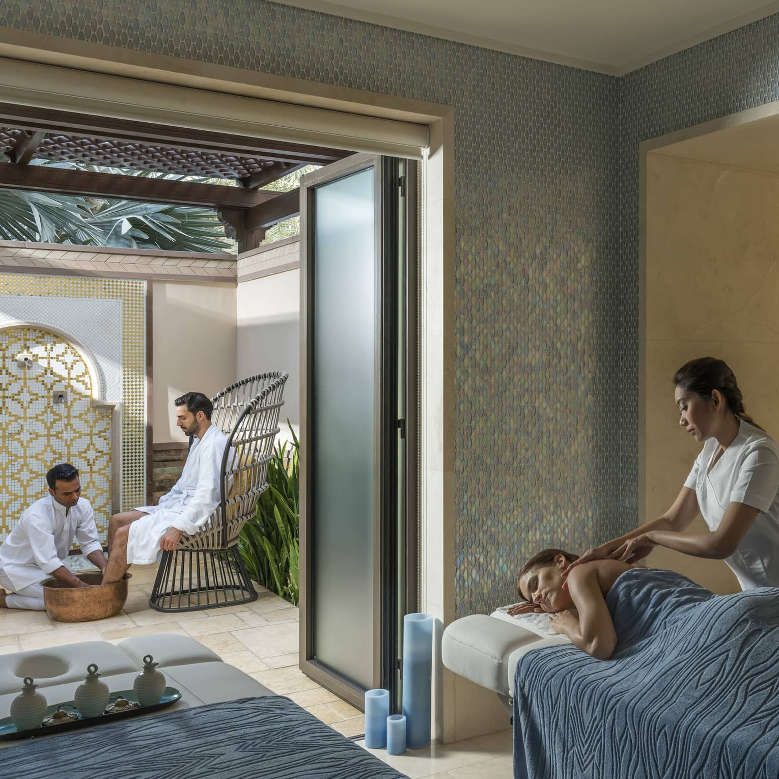 A woman under a blue sheet gets a massage from a masseuse surrounded by candles and natural light. 