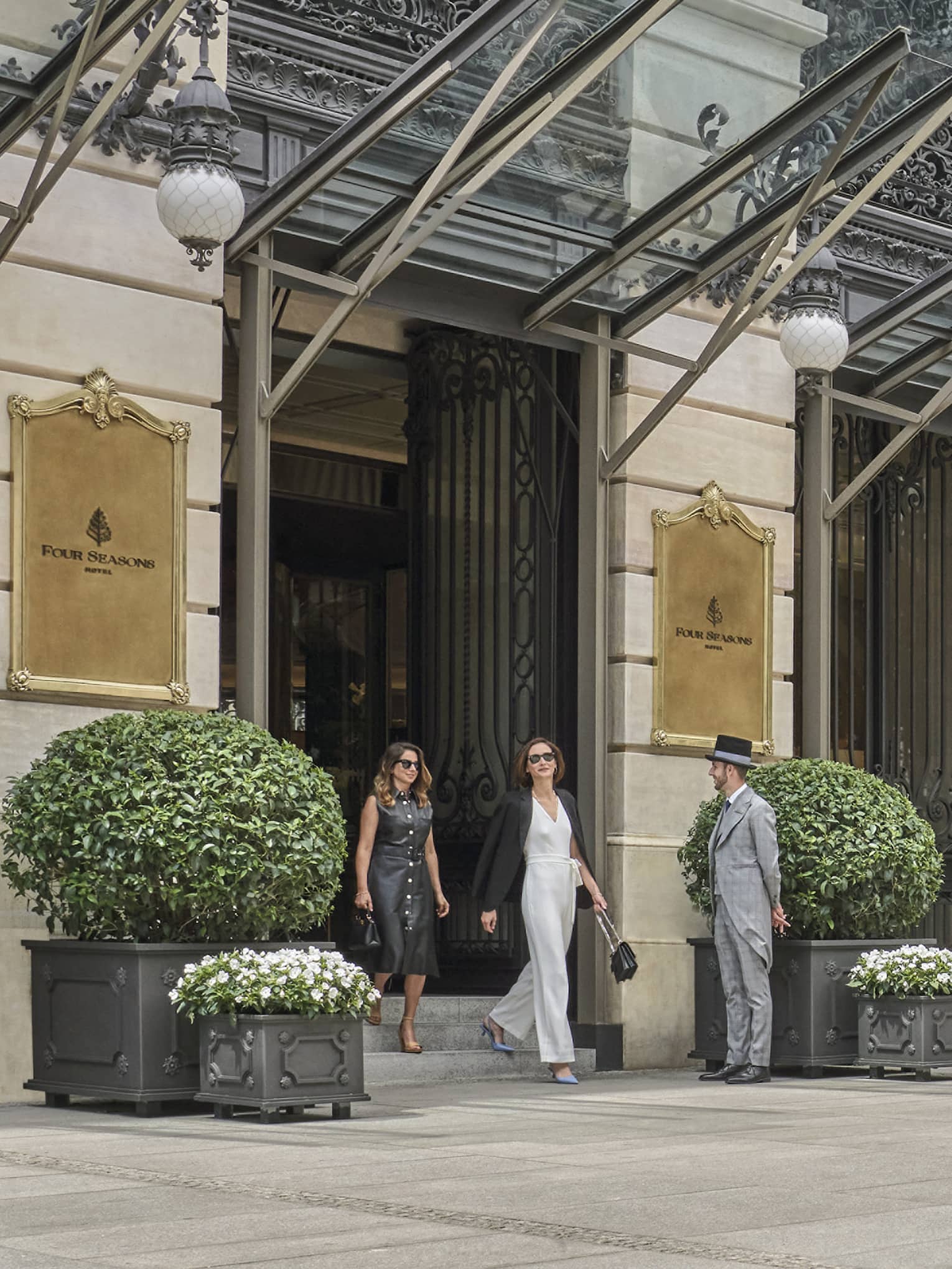 Four Seasons luxury hotel entrance with two guests walking out, doorman, glass canopies and large potted plants