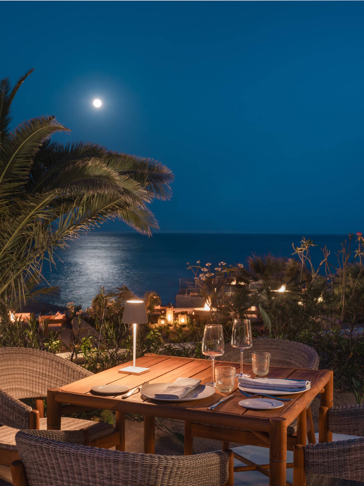 Dining table set for four on an outdoor patio at night with palm trees and the ocean in the distance