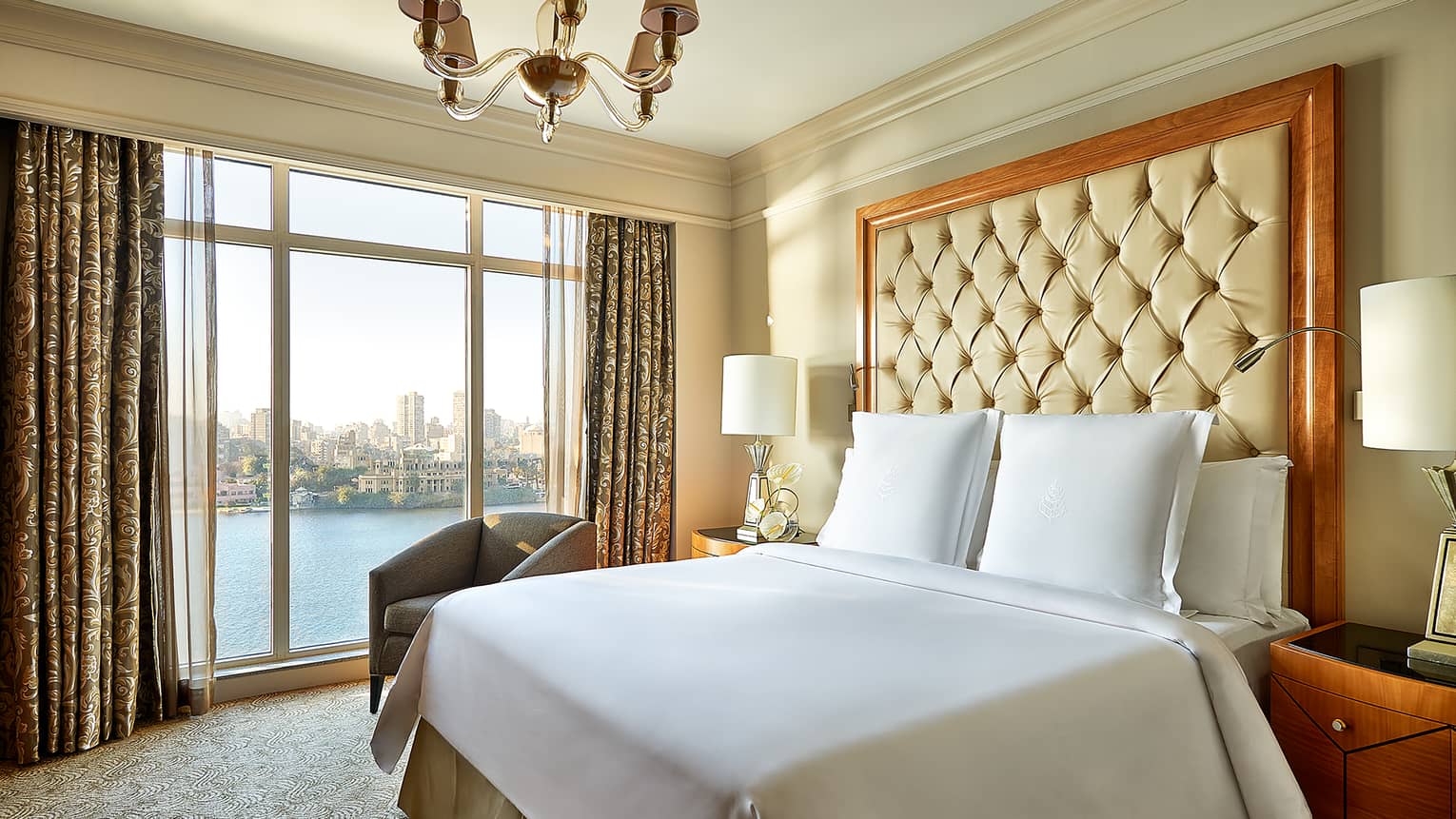 Diplomatic Suite bed with white bedspread, pillows and tall beige leather headboard beside tall window with river view