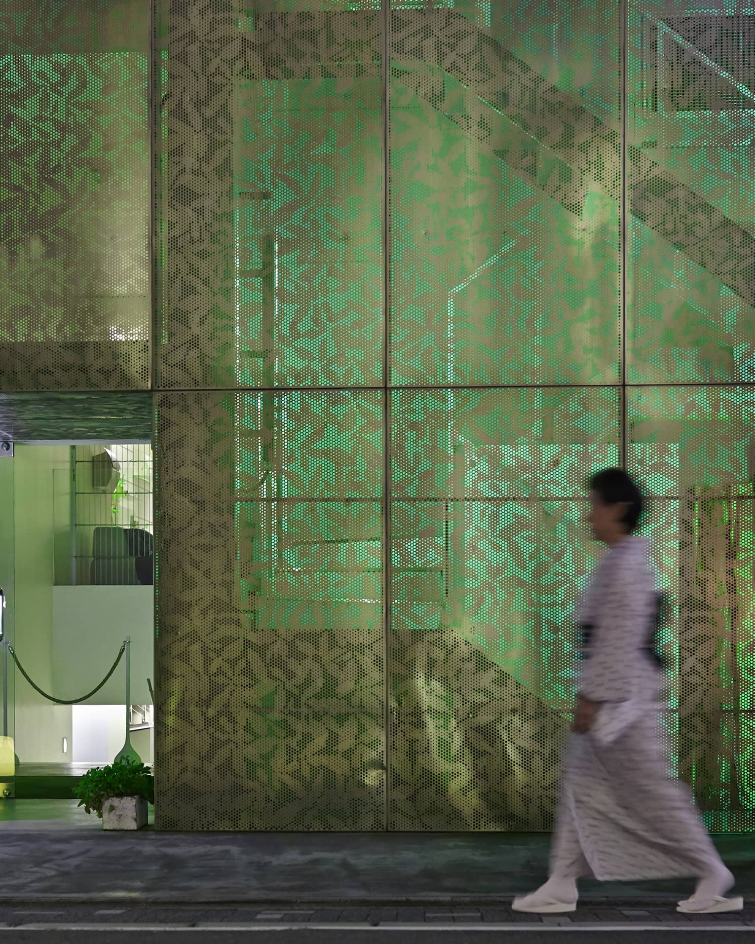 A person heads toward a door to a modern room, passing a multi-paned screened wall with a leaf pattern illuminated in green.