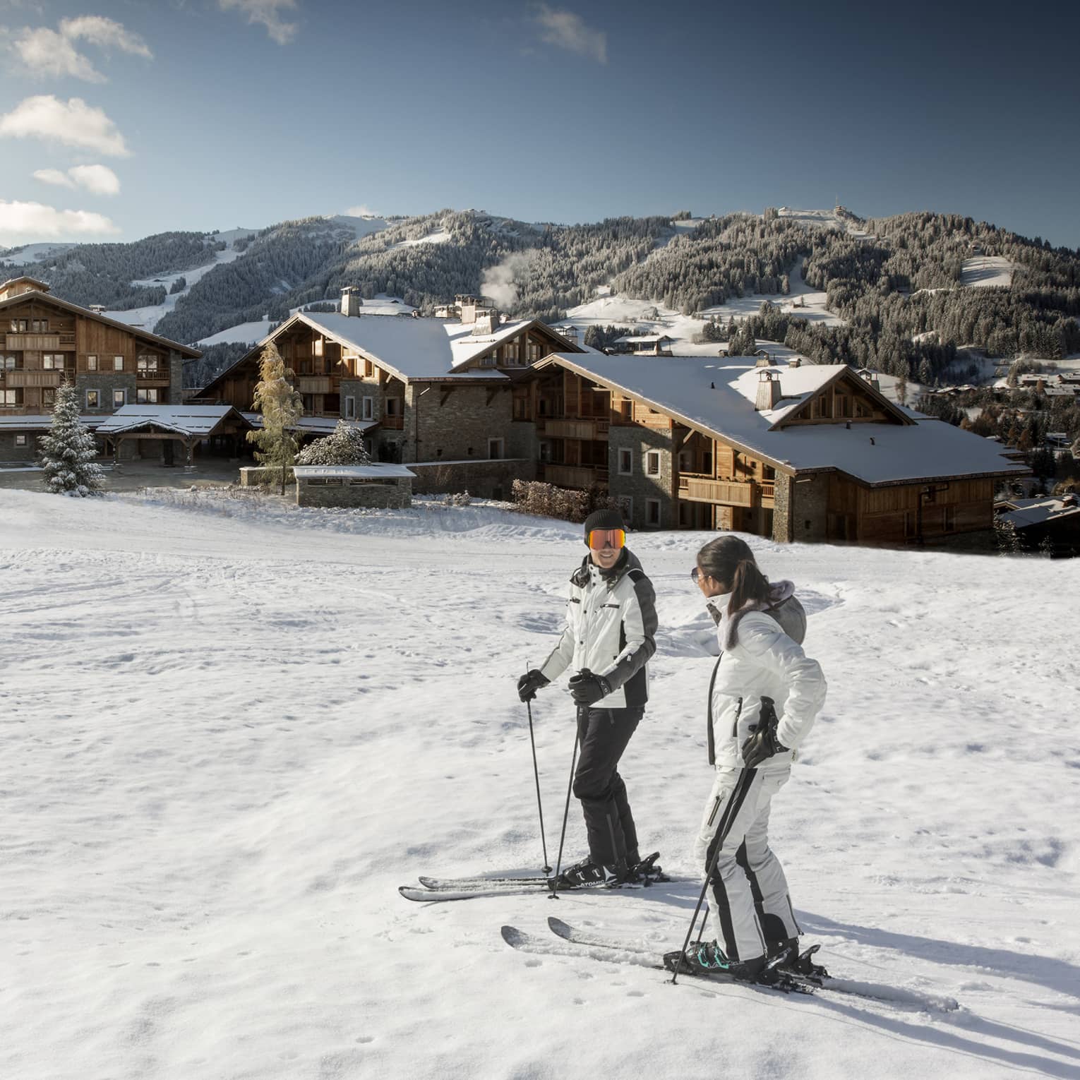 Man, woman on skis at top of snowy hill, Four Seasons Hotel Megeve in background