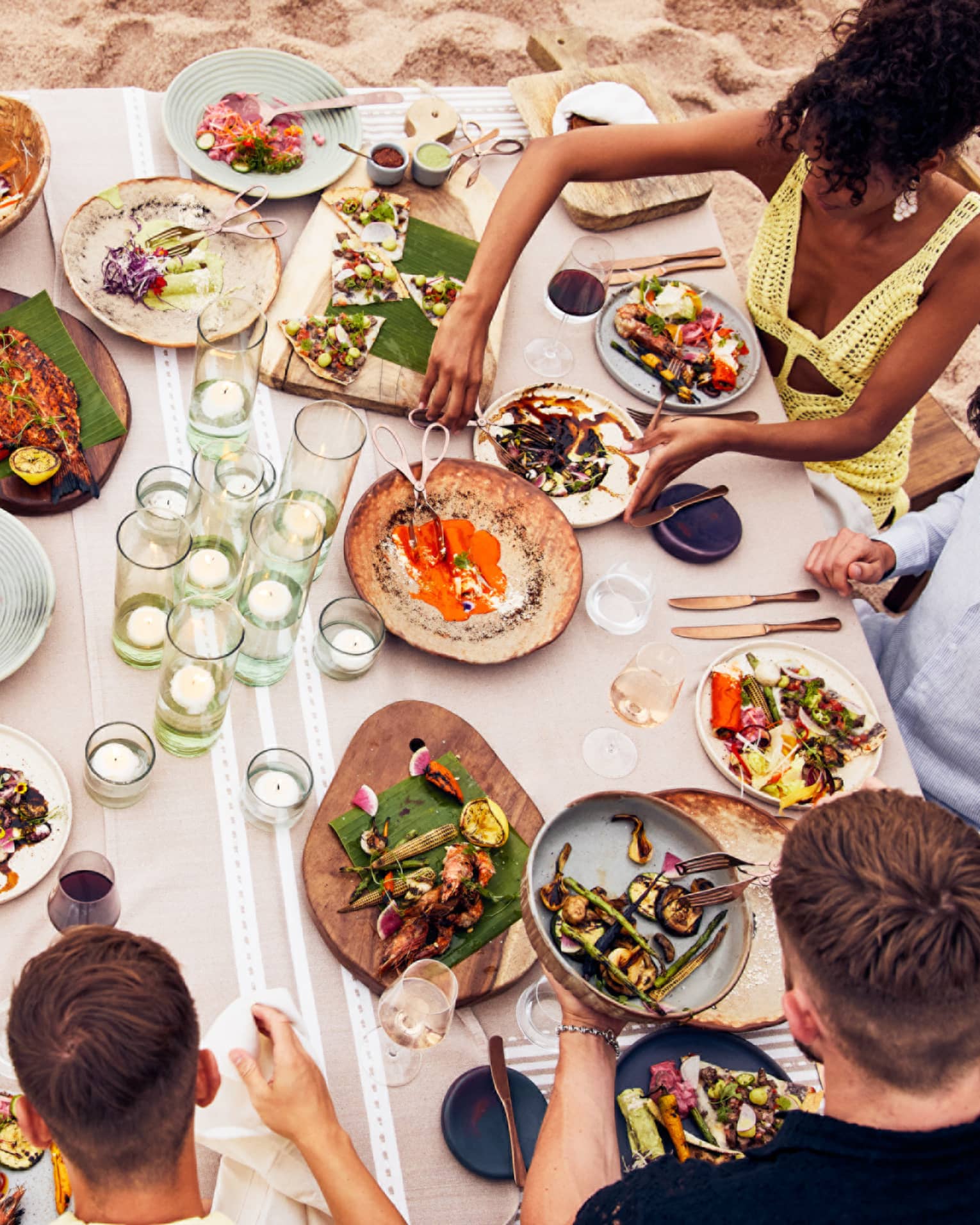 Beachside dining table with an assortment of vibrant dishes, candlelit glass holders and guests enjoying fresh seafood and grilled vegetables