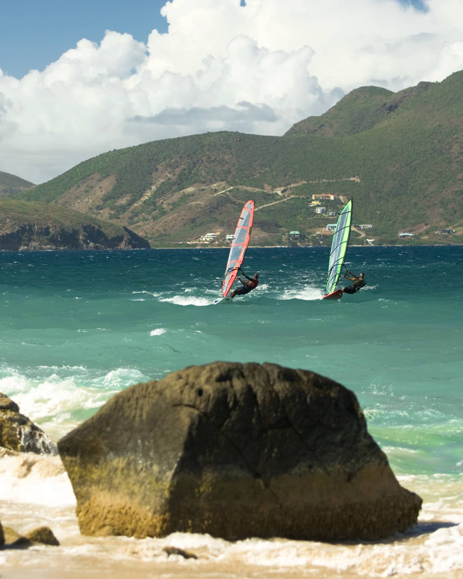 Two windsurfers glide on waves in bay between rocks, green mountains
