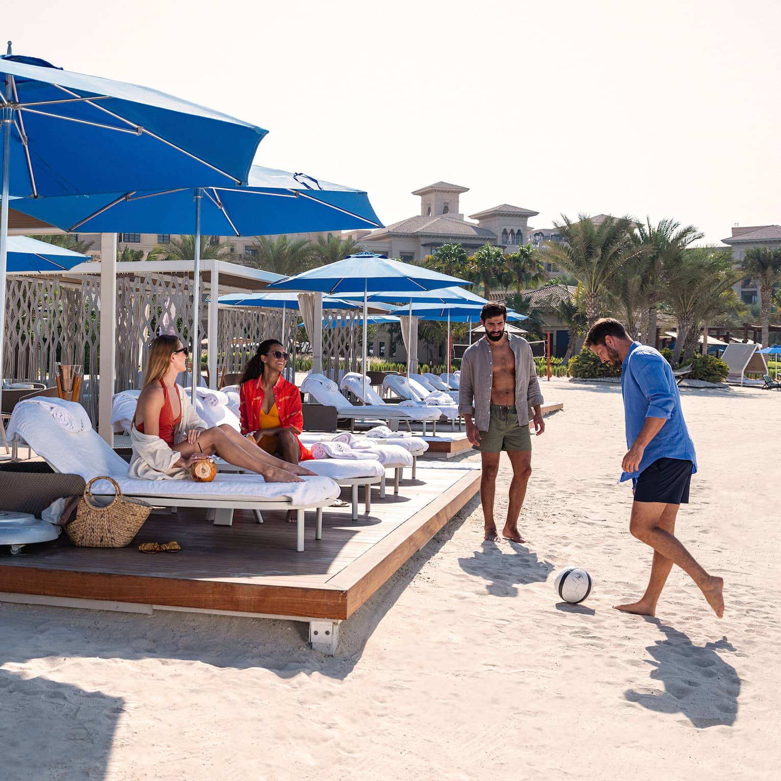 Two beachgoers play soccer while two others sit on lounge chairs, sheltered under blue umbrellas on a beach cabana platform.