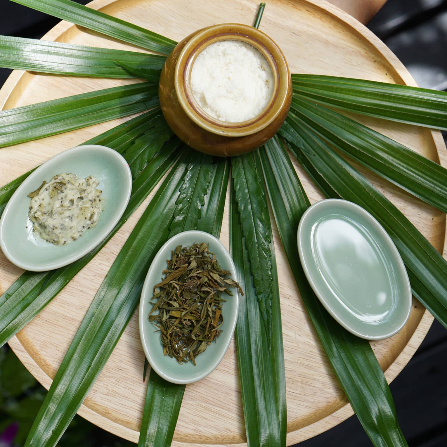 Pandan leaves laid out like a fan on a wooden table with a jar and small bowls of herbs and scrubs placed on top.