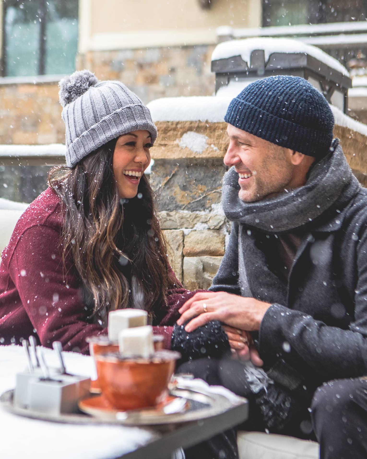 Smiling couple sitting by a firepit in a snowy outdoor setting, bundled in winter coats and hats. Snowflakes fall around them as they enjoy a cosy moment. 