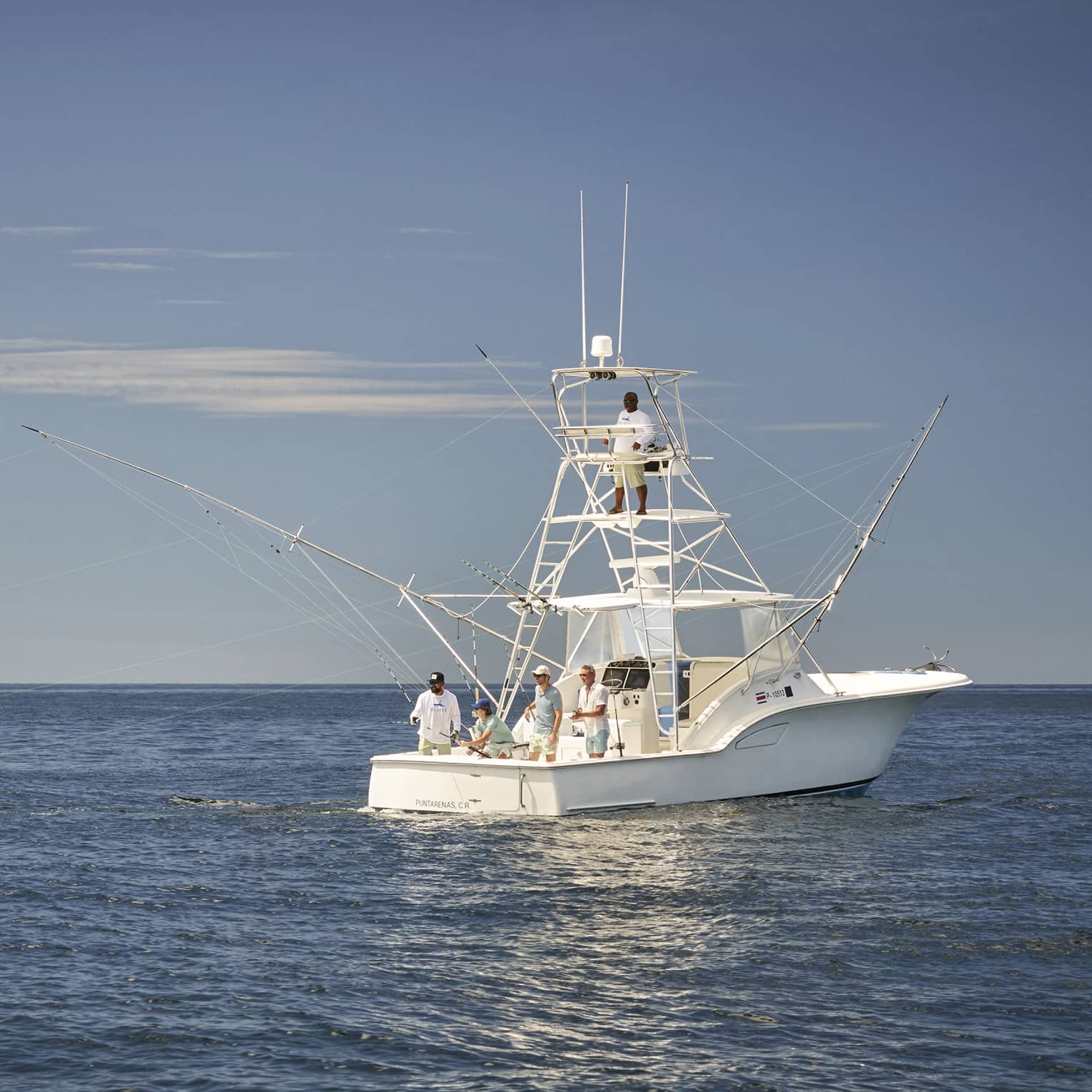 Long view of four guests on a fishing boat, a guide perched on a high platform lookout, on deep-blue water under a clear sky.