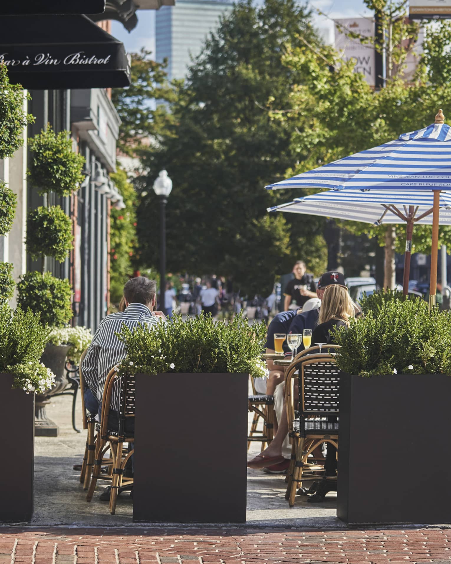 People dine on Bar a Vin Bistrot sidewalk patio behind planters, umbrellas