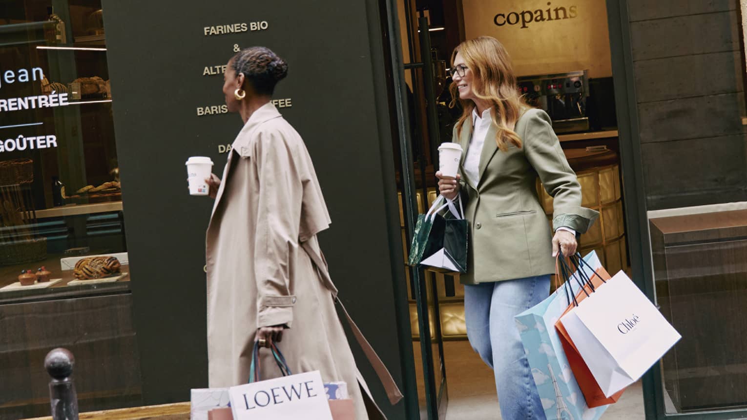 Fashionably dressed tourists with coffee and shopping bags exit an upscale café. Pastries are displayed in the café window.
