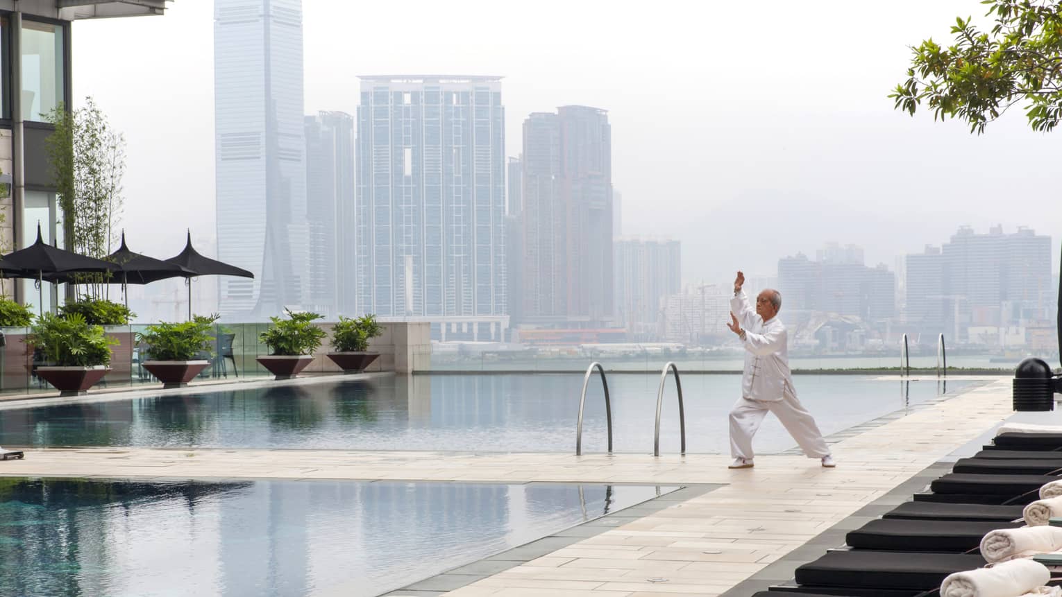 Tai Chi master wearing white holds pose on patio deck by swimming pools, misty skyline