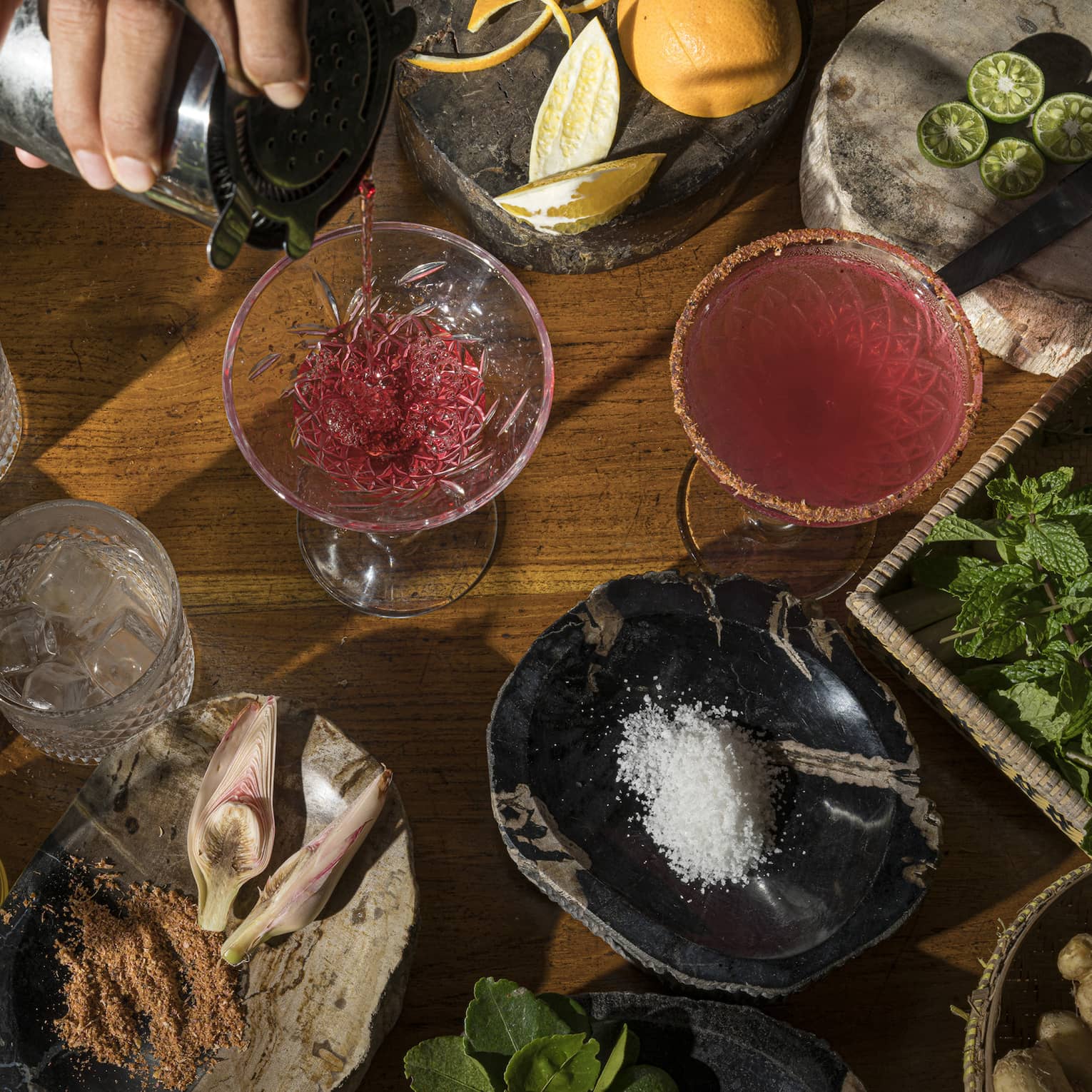 Aerial view of bartender's hand pouring ruby-coloured cocktail intro stemmed glass, local ingredients on wooden bartop surrounding it